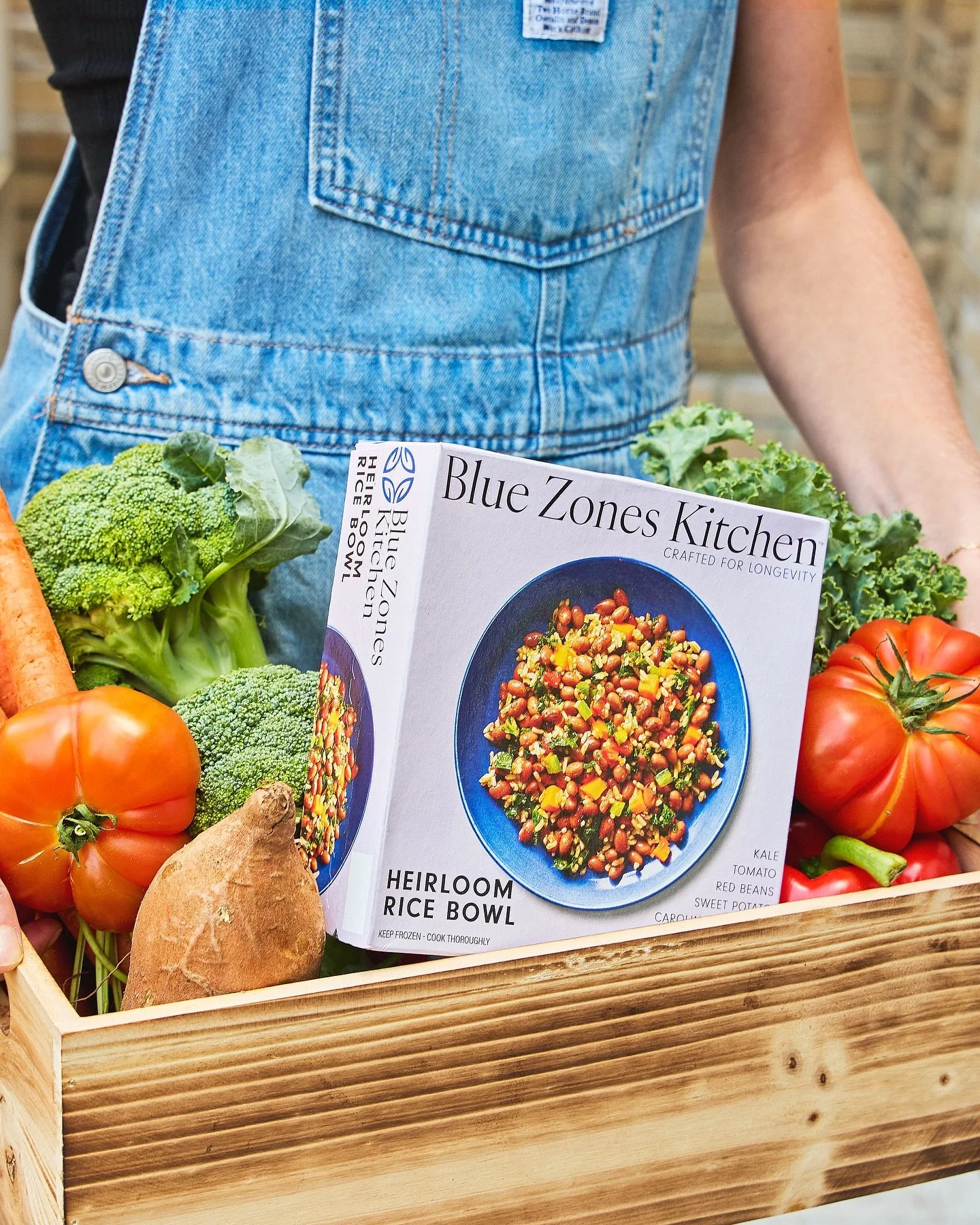 A wooden crate filled with fresh vegetables and a magazine about heirloom rice bowl recipes. The vegetables include broccoli, carrots, sweet potatoes, red tomatoes, and kale. The magazine features a colorful rice dish on a blue plate, titled "Blue Zones Kitchen".
