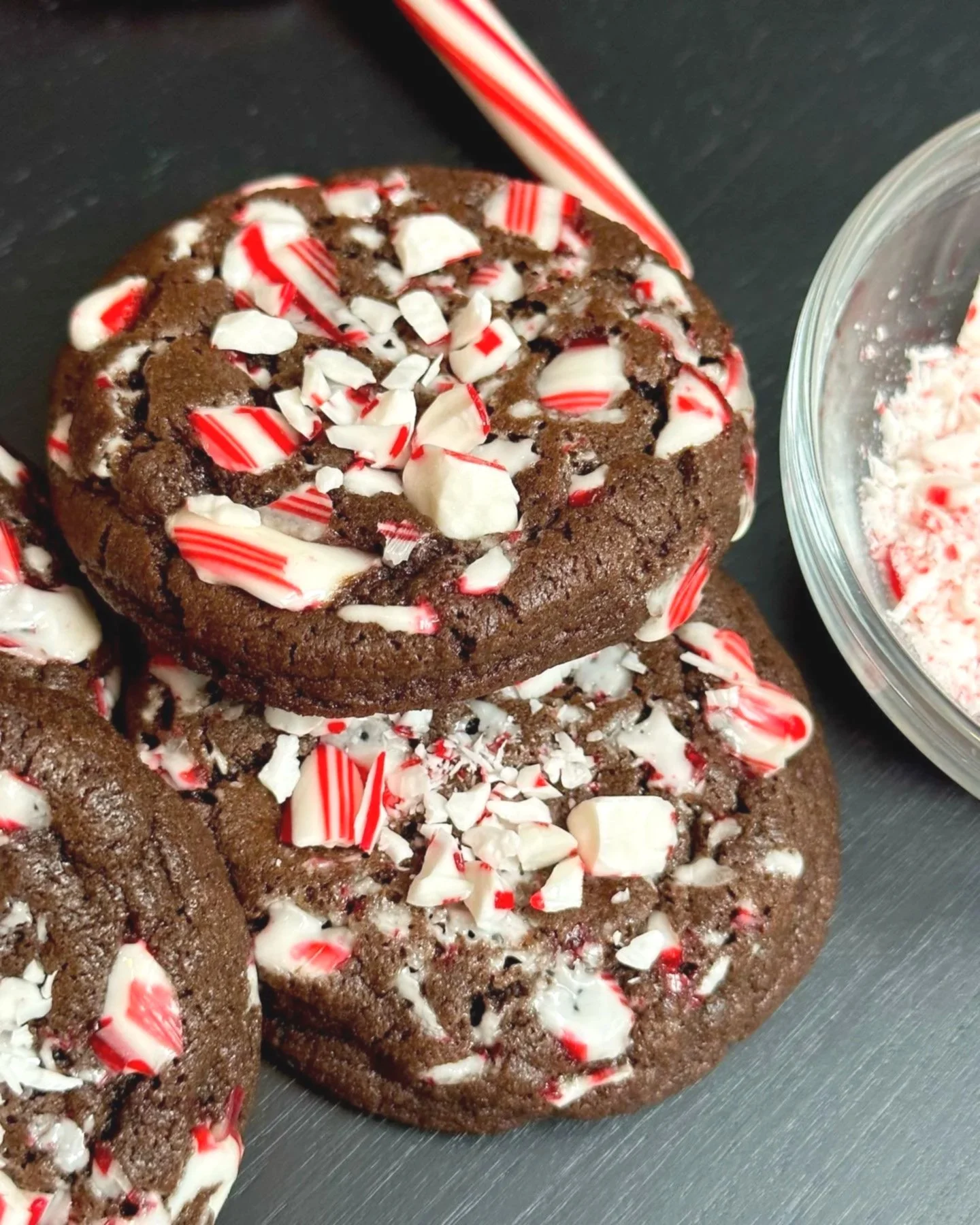 Chocolate cookies with crushed peppermint candies on top, with a candy cane and a bowl of crushed peppermint in the background.