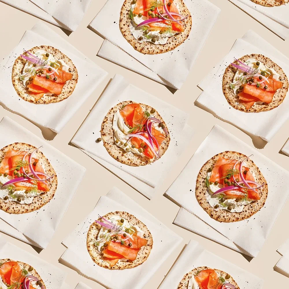 Multiple individual pizzas topped with smoked salmon, cream cheese, red onions, capers, and herbs arranged on white surfaces.