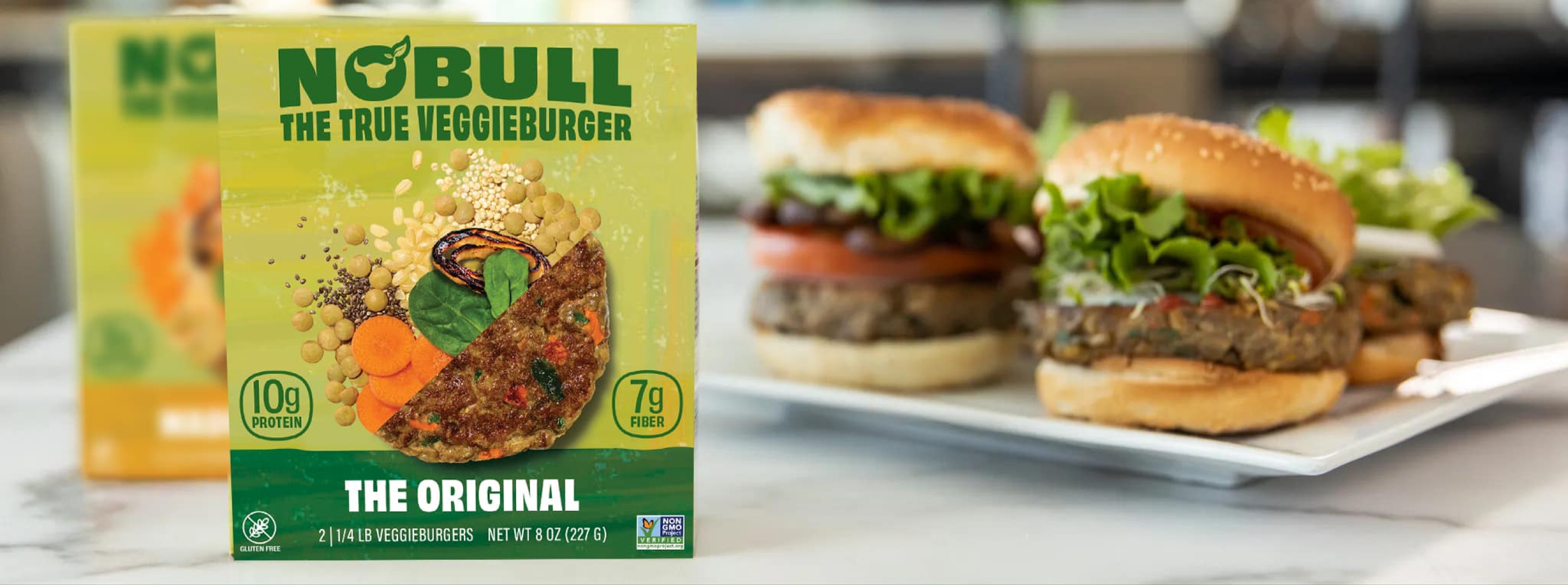 Box of Nobull veggie burgers next to two prepared veggie burger sandwiches with lettuce, tomato, and veggie patty on a white plate.
