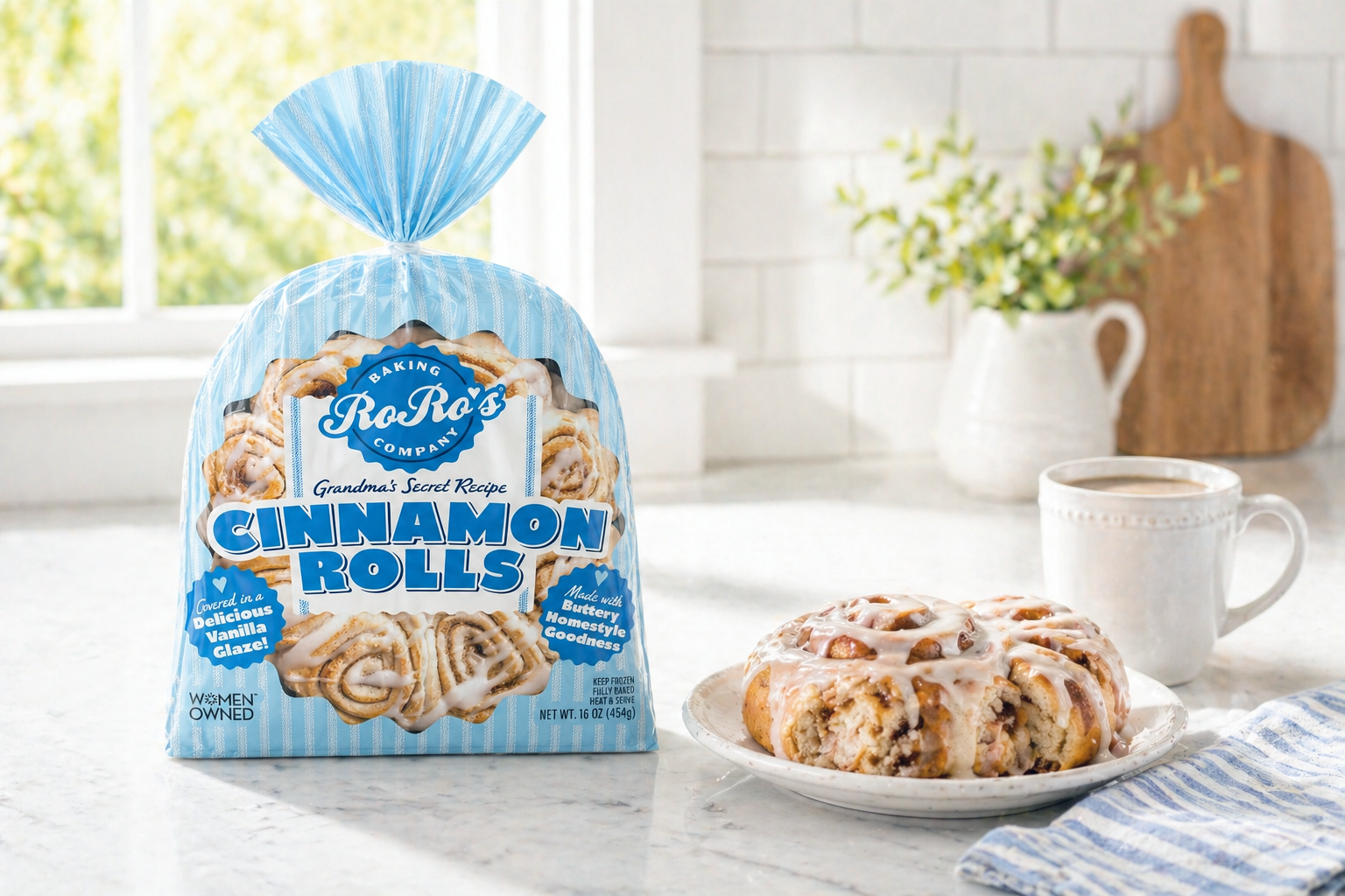 A bag of Ra Ro's Cinnamon Rolls on a kitchen counter next to a plate with a cinnamon roll topped with glaze, a white mug of coffee, a vase with flowers, and a cutting board in the background.