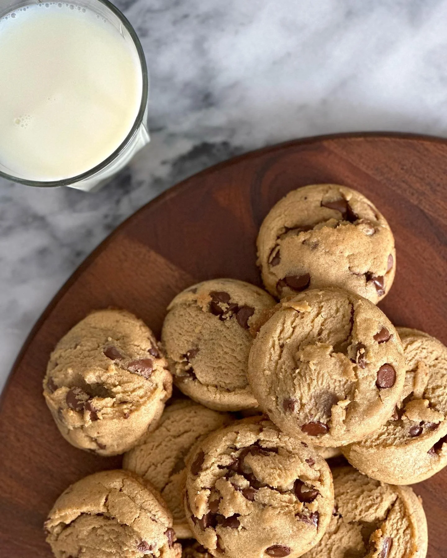 A wooden plate with several chocolate chip cookies, and a glass of milk on a marble countertop.