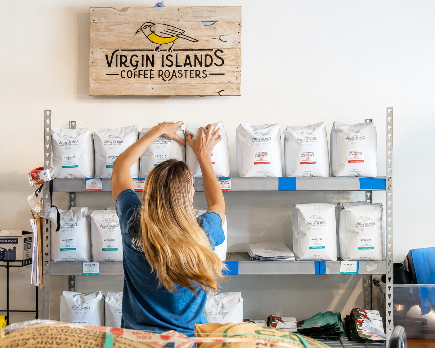 A woman with long blonde hair stocking Virgin Islands Coffee Roasters bags on metal shelves inside the store.