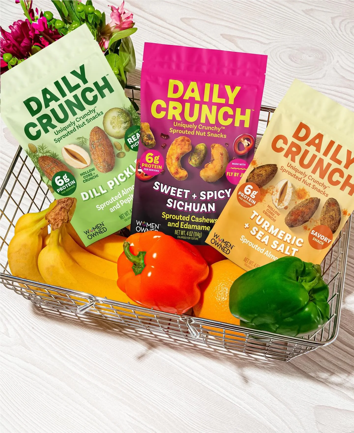 A shopping basket filled with colorful bell peppers, bananas, and tomatoes, with three bags of Daily Crunch sprouted nut snacks standing behind the vegetables.