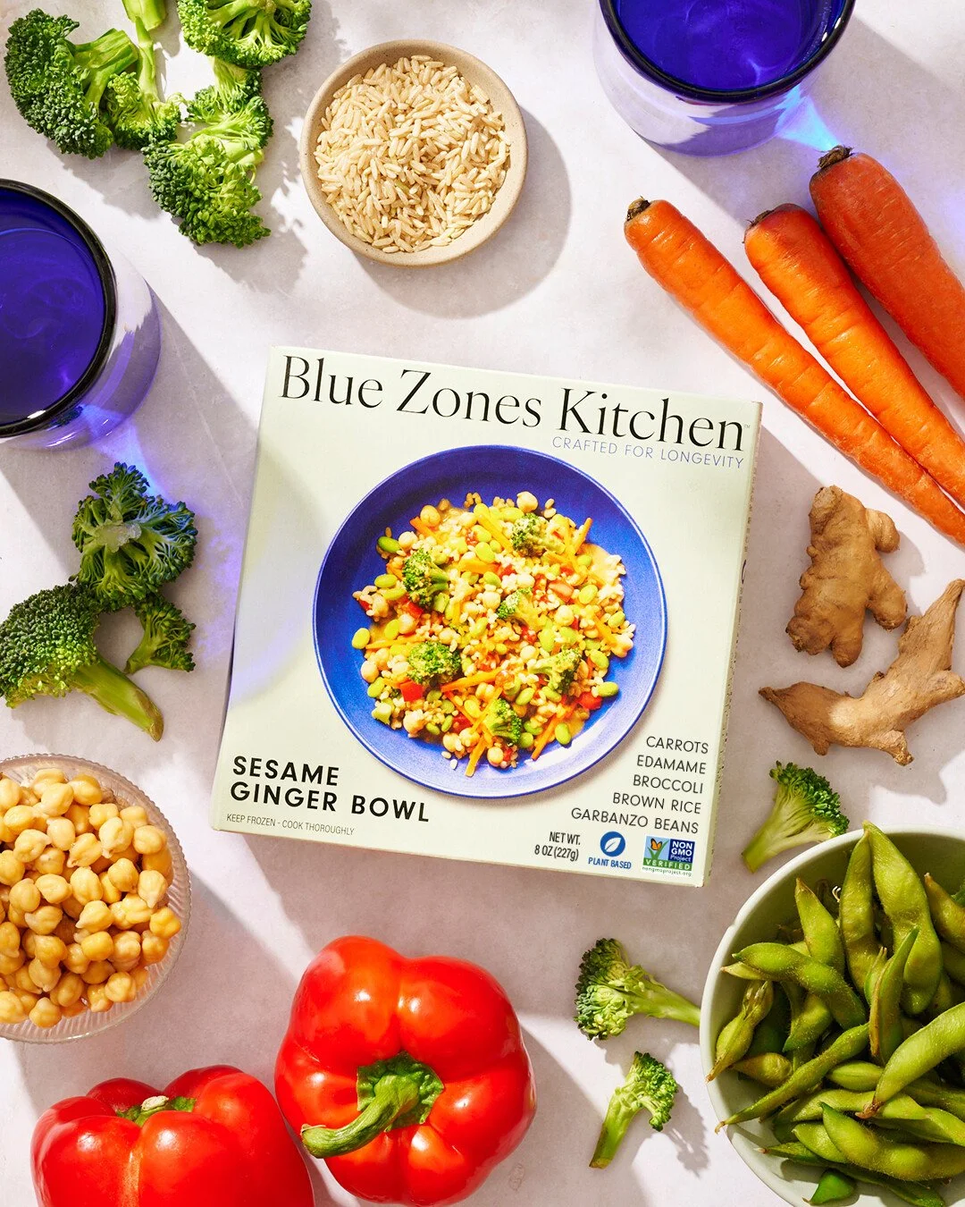 A variety of fresh vegetables including broccoli, carrots, ginger, red bell peppers, and edamame are arranged around a boxed frozen vegetable blend labeled 'Blue Zones Kitchen Sesame Ginger Bowl,' with two glasses of blue beverage and a bowl of rice also visible.