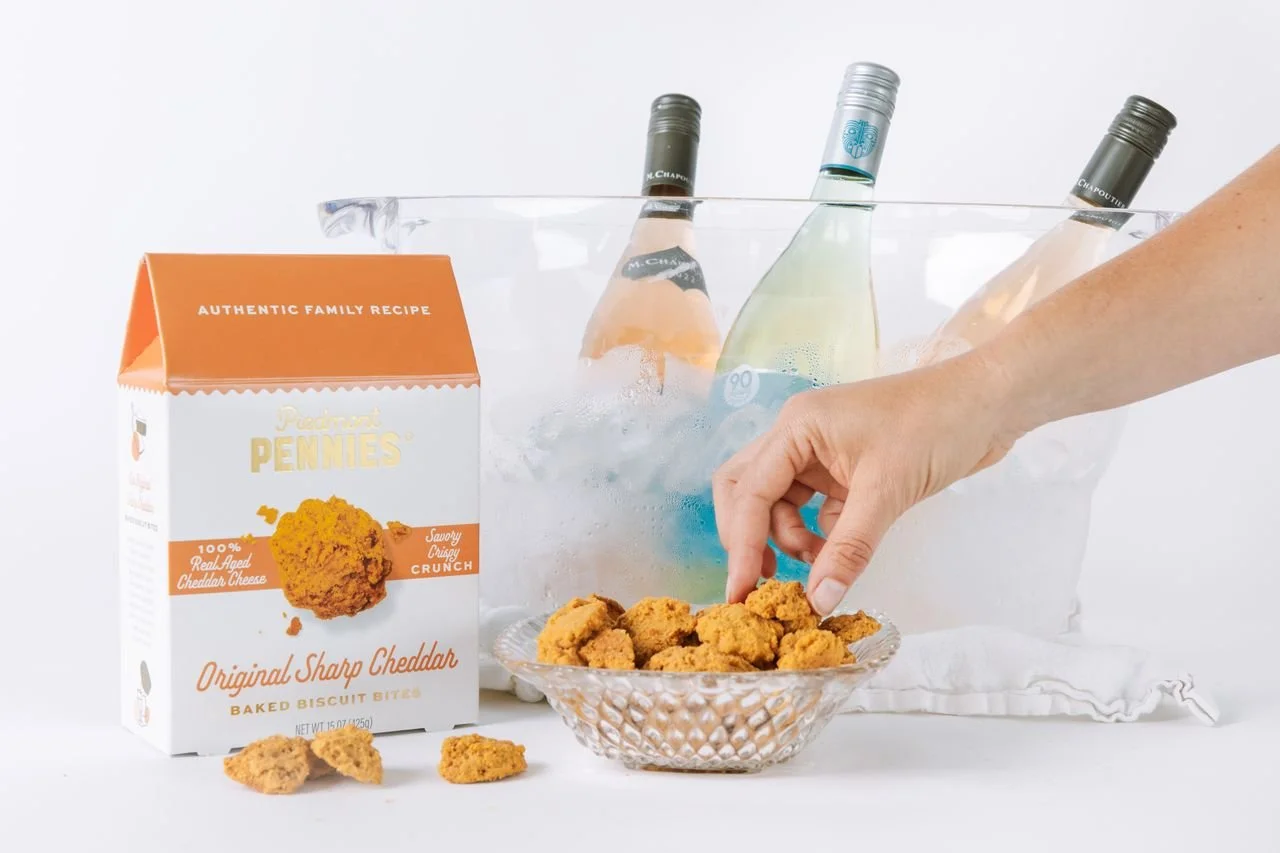 A person's hand reaching for cookies in a glass bowl, with a box of snack crackers and bottled drinks in ice in the background.