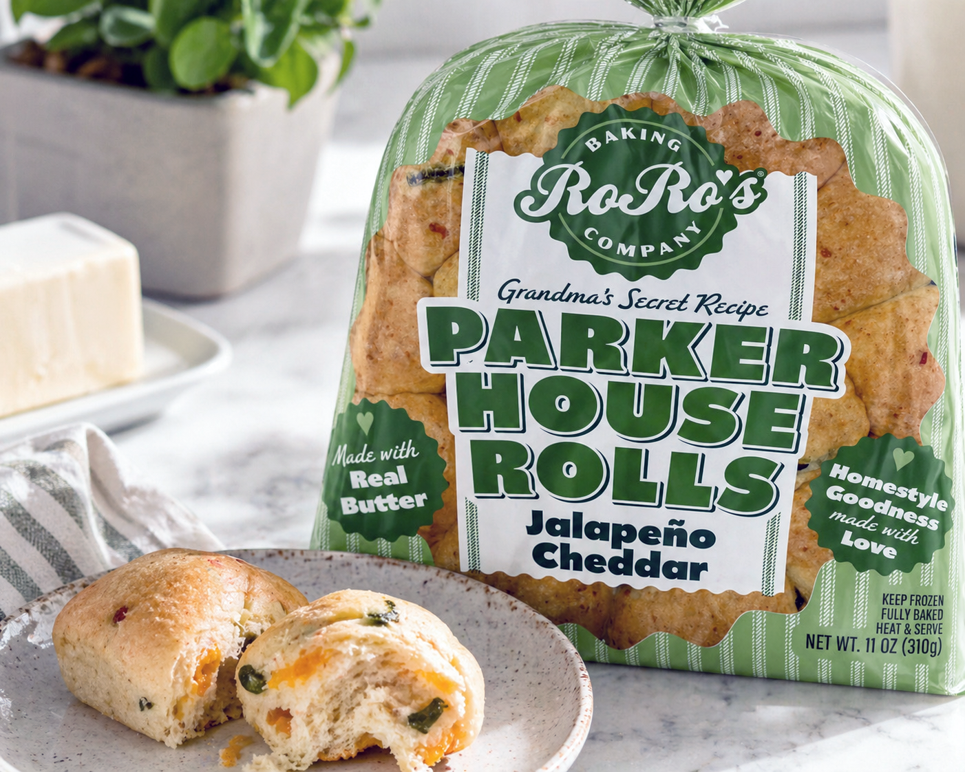 Package of Ro Ro's Grandma's Secret Recipe Parker House Rolls with Jalapeño Cheddar, with some rolls on a plate and a block of cheese in the background.
