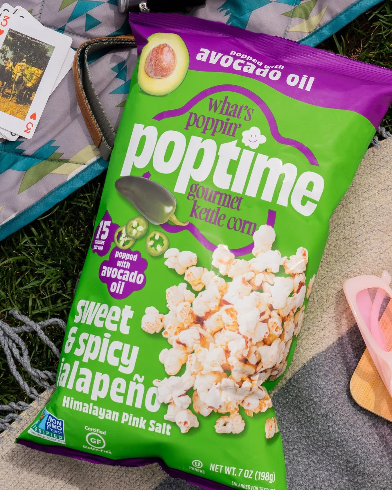 Bag of sweet and spicy Himalayan pink salt flavored popcorn with avocado oil, placed on a grassy surface next to playing cards and a gray cloth.