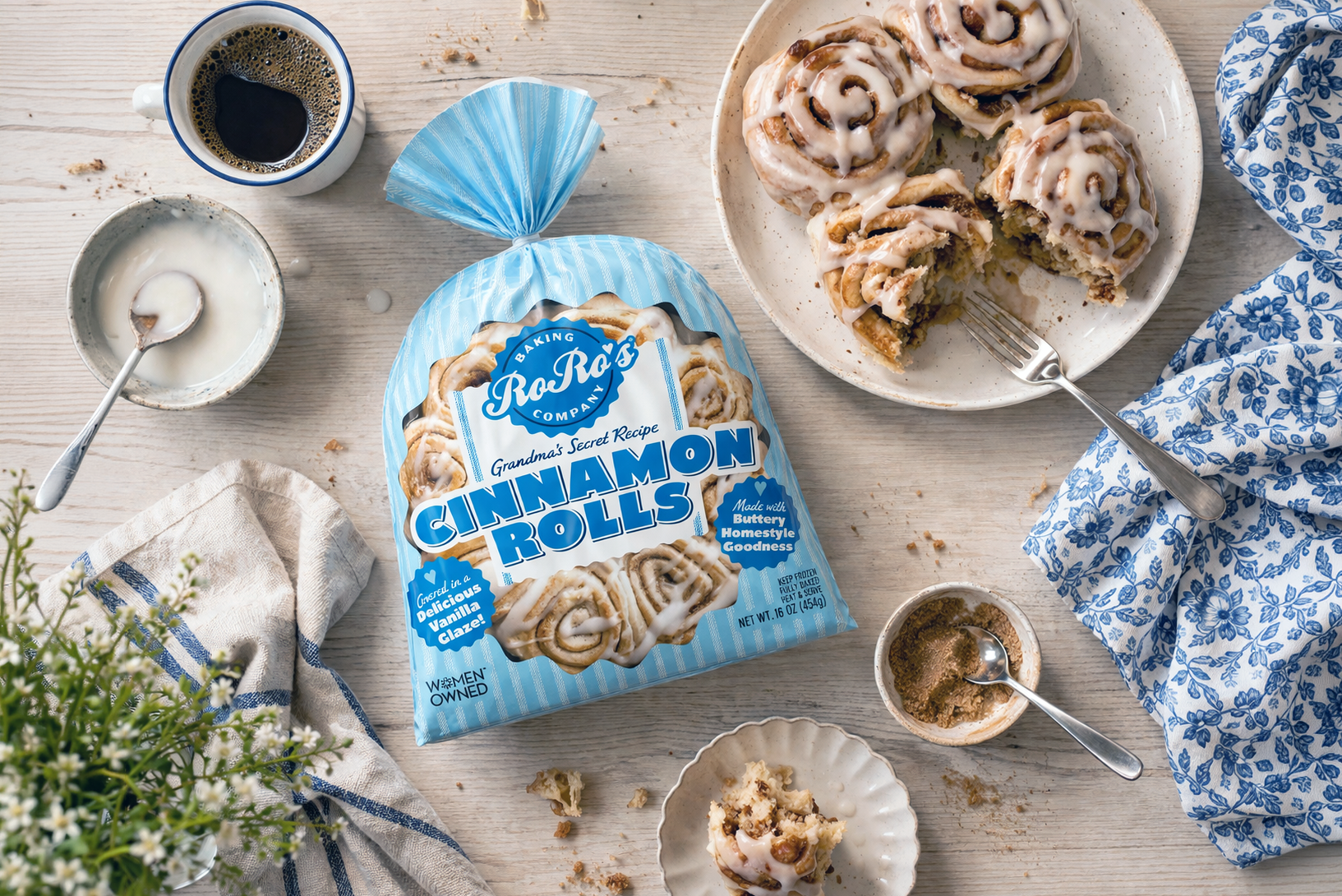 A white wooden table with cinnamon rolls topped with glaze, a cup of black coffee, a bowl of icing, a small bowl with cinnamon sugar, a packet of cinnamon rolls, blue patterned napkins, and some scattered crumbs and flowers.
