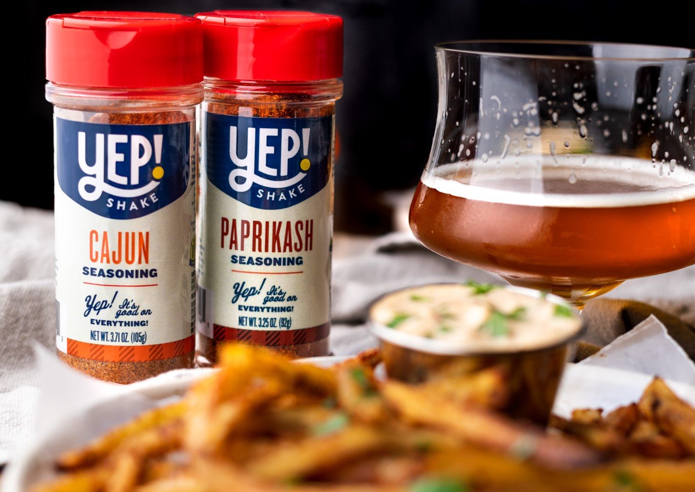 Two bottles of YEP! Shake seasoning, one Cajun and one Paprikash, placed on a table alongside a glass of beer, a bowl of creamy dip garnished with herbs, and a dish of fried food, all with a dark background.
