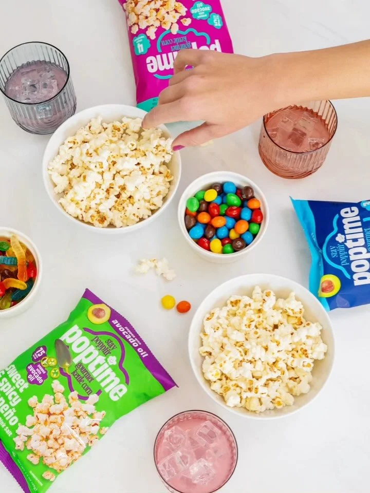 A table with snacks including two bowls of popcorn, a bowl of colorful candy-coated chocolates, a bag of Pop-Tarts, and a bag of Popcorn. Two glasses with pink-colored drinks and a hand reaching for popcorn are also on the table.