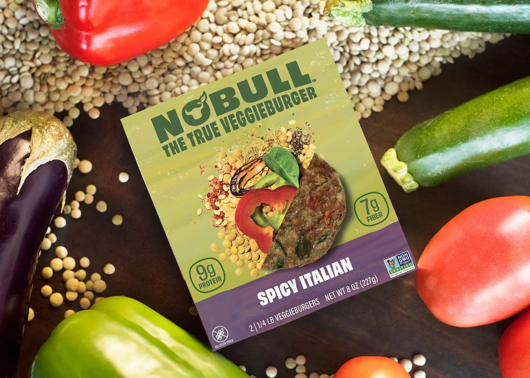 Packaged veggie burger with label reading "No Bull, The True Veggieburger, Spicy Italian" surrounded by fresh vegetables including tomato, cucumber, eggplant, and green bell pepper on a dark surface.