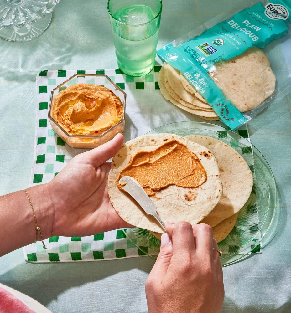 Person spreads peanut butter on a tortilla with a butter knife, with more tortillas and peanut butter on a plate, a glass of water, and a pack of tortillas on the table.