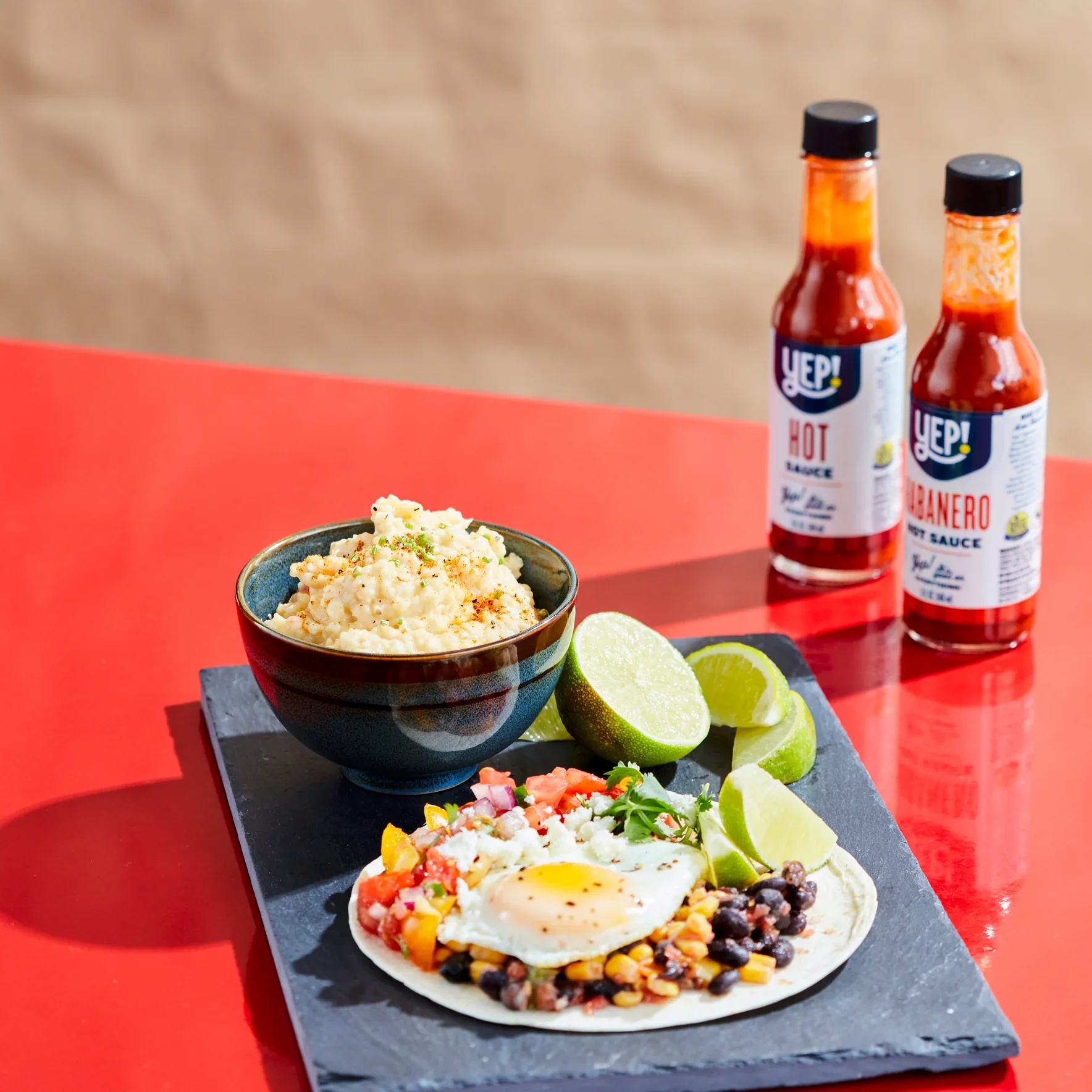 A meal plate with a breakfast, topped with a fried egg, accompanied by lime wedges, and small bowls of mashed potatoes and salsa on a black slate platter, with two bottles of hot sauce in the background.