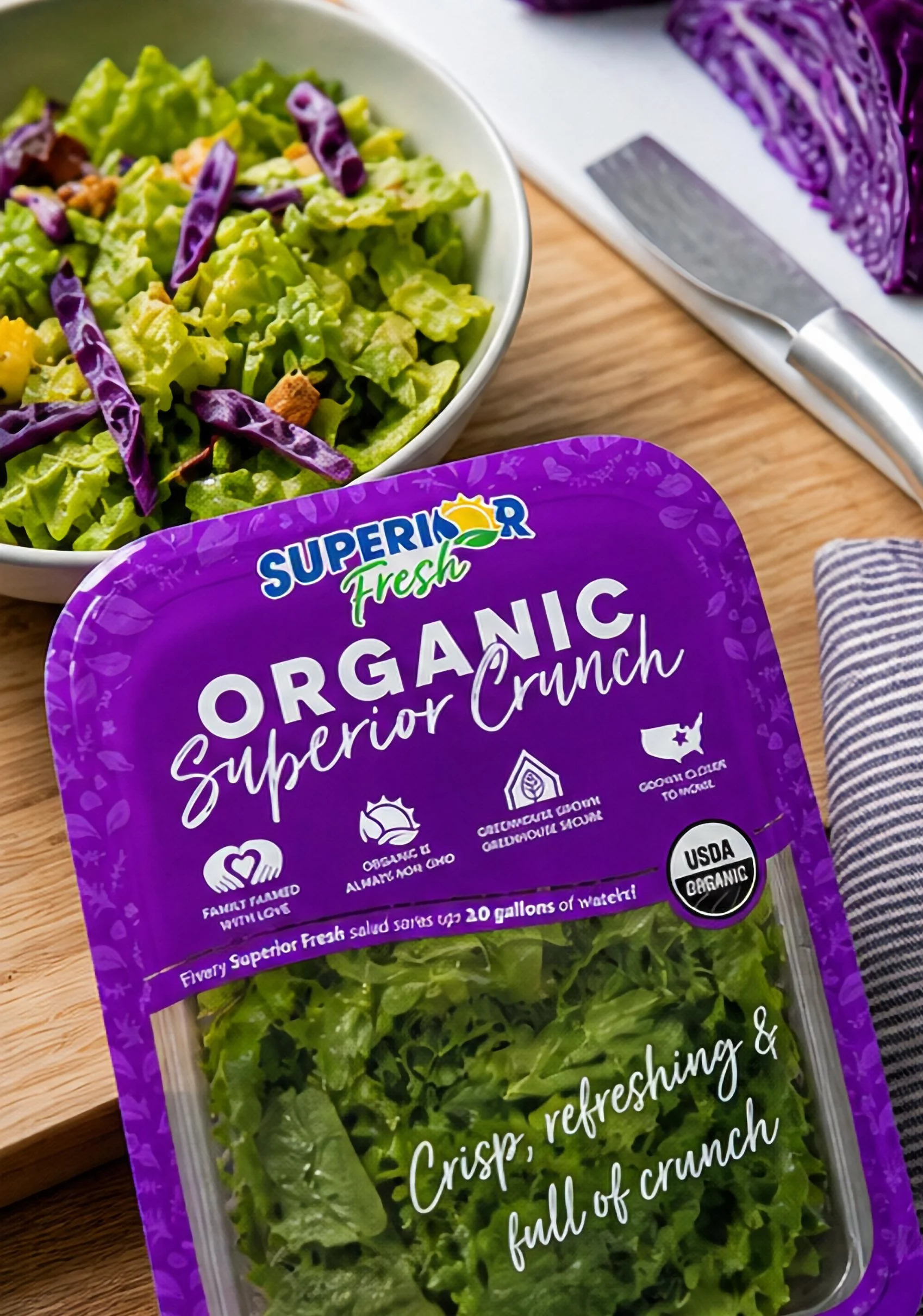 Fresh organic arugula salad in a plastic container with purple packaging from SuperRior Organic. Sliced purple cabbage on a cutting board nearby, a steak knife resting on a white cloth.