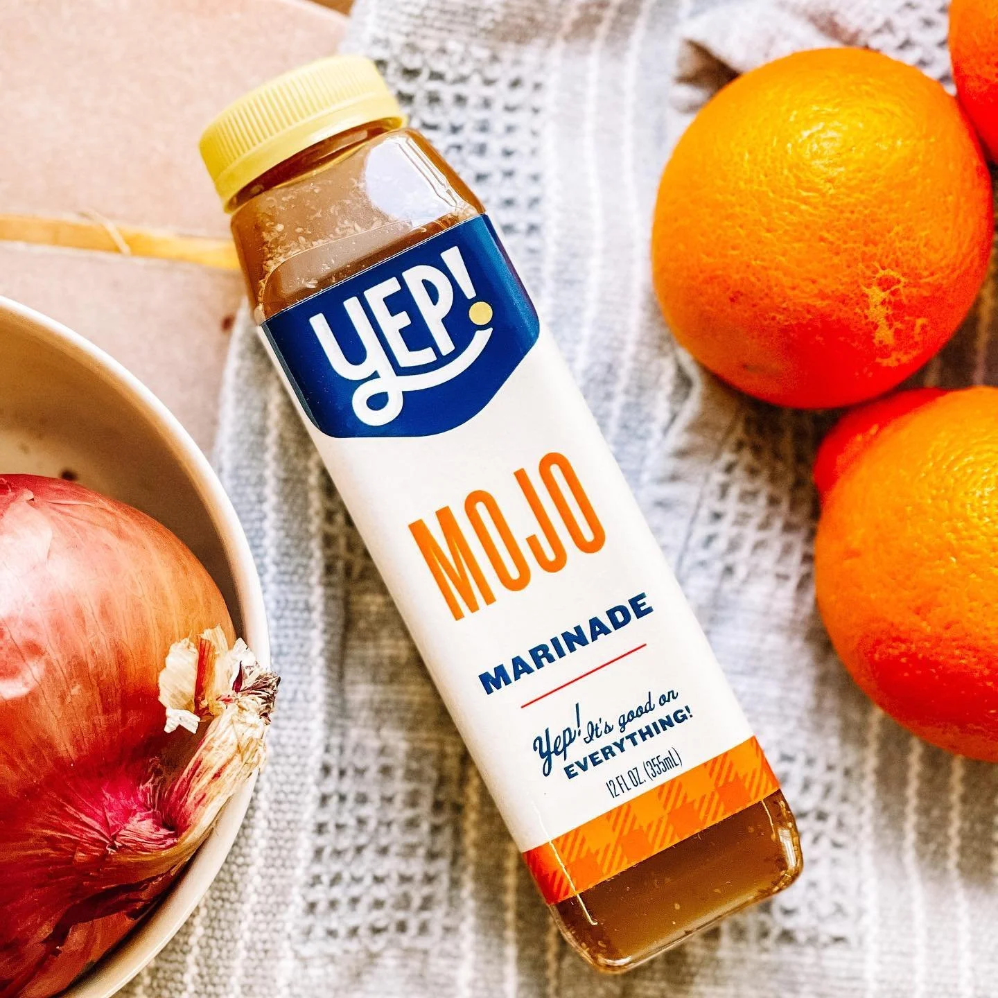 A bottle of mojo marinade surrounded by oranges, onions, and a towel.