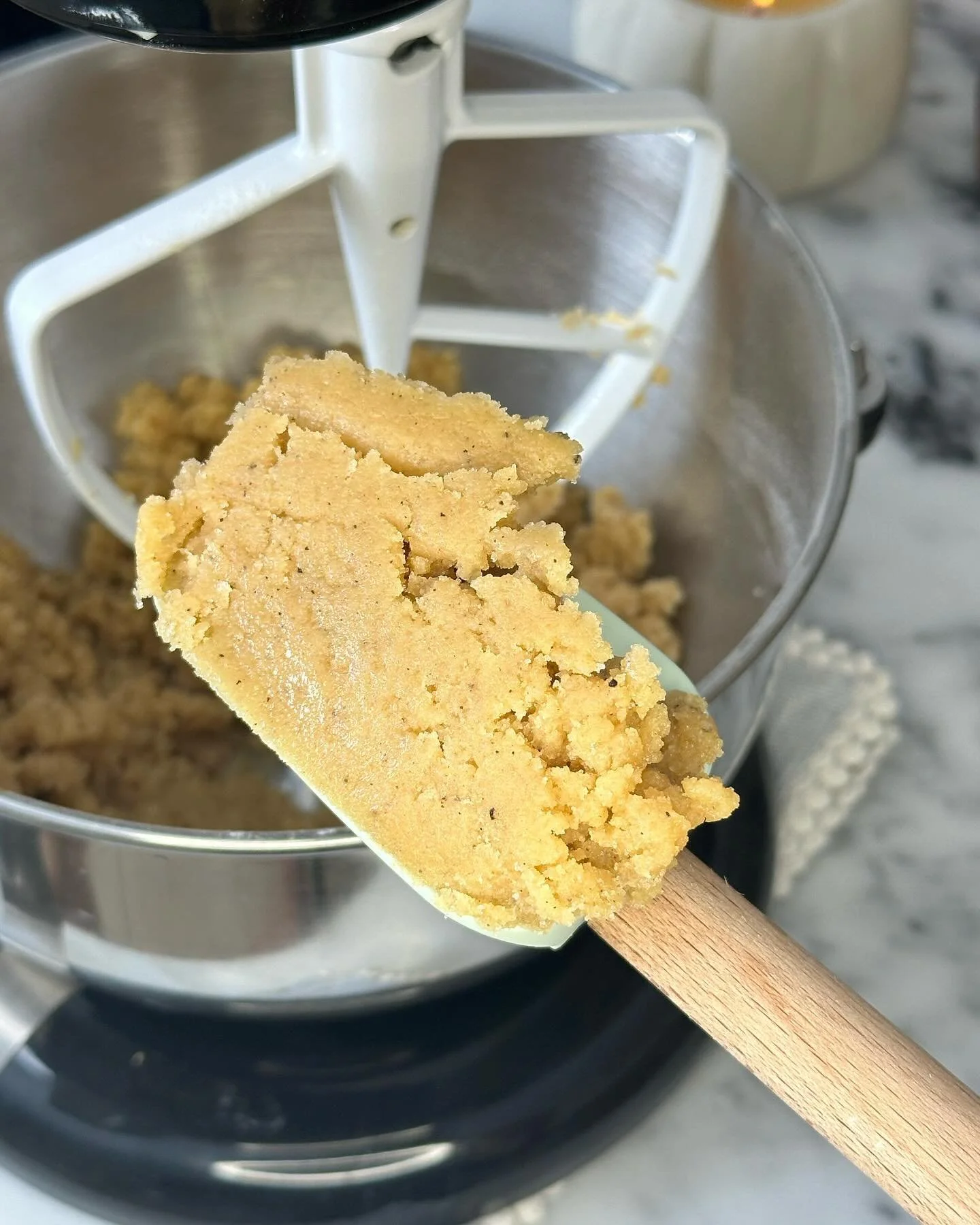 A close-up of a spatula with cookie dough, being used in a stand mixer with a metal bowl, in a kitchen setting.