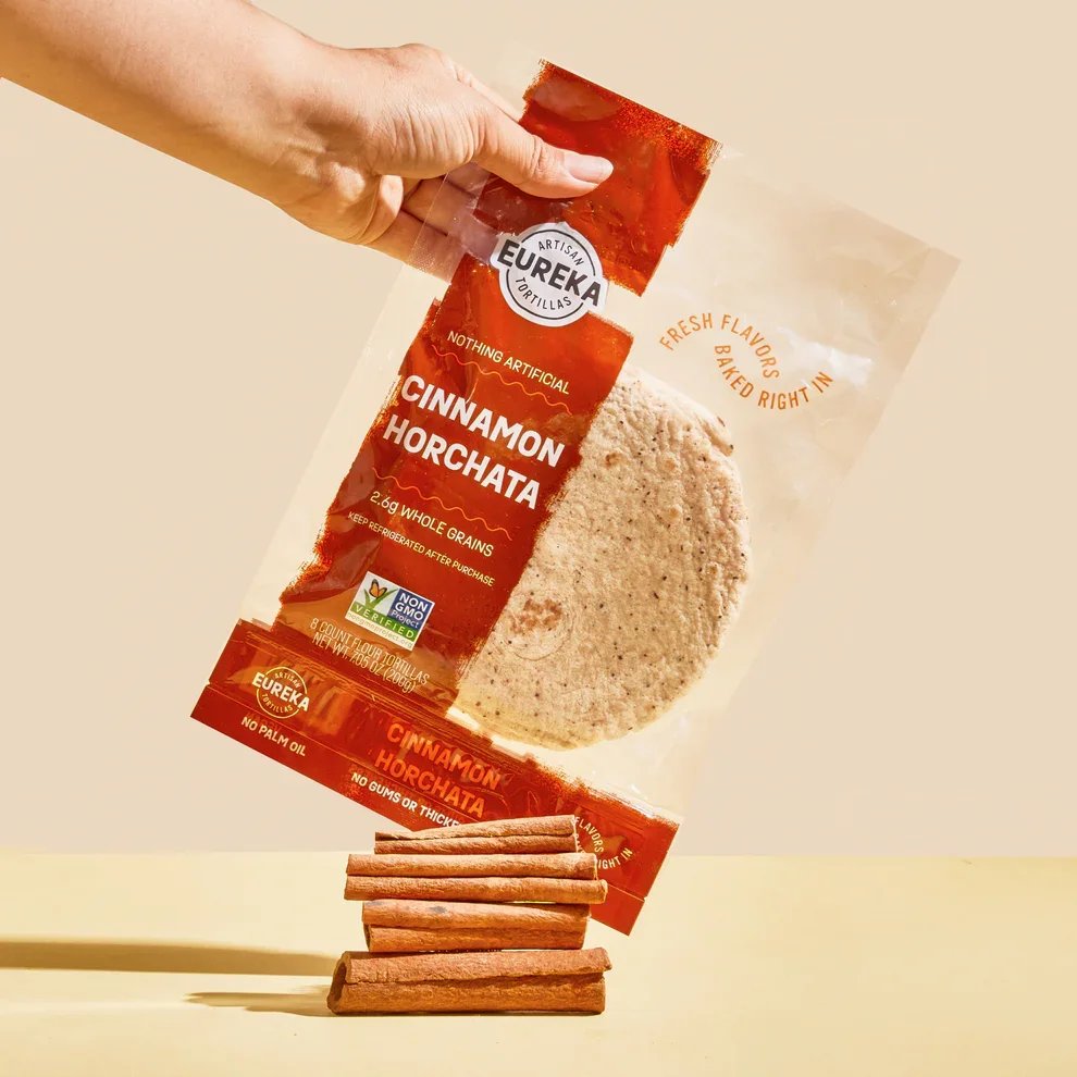 A hand holding a cinnamon horchata flavored tortilla chip in front of a package of Eureka Cinnamon Horchata tortillas, with cinnamon sticks placed on a flat surface below.