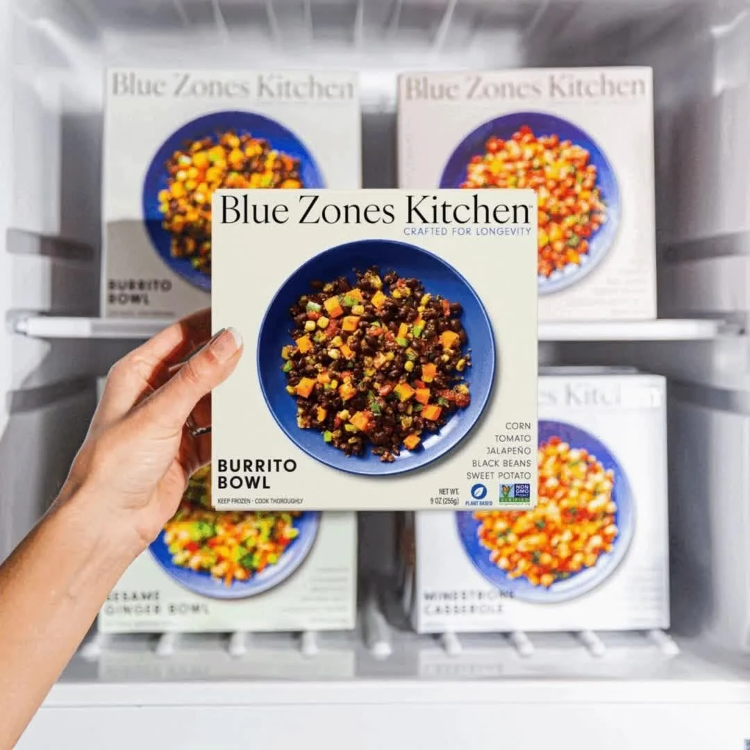 A hand holding a boxed frozen burrito bowl from Blue Zones Kitchen in front of a refrigerated shelf stocked with other boxes of the same product.
