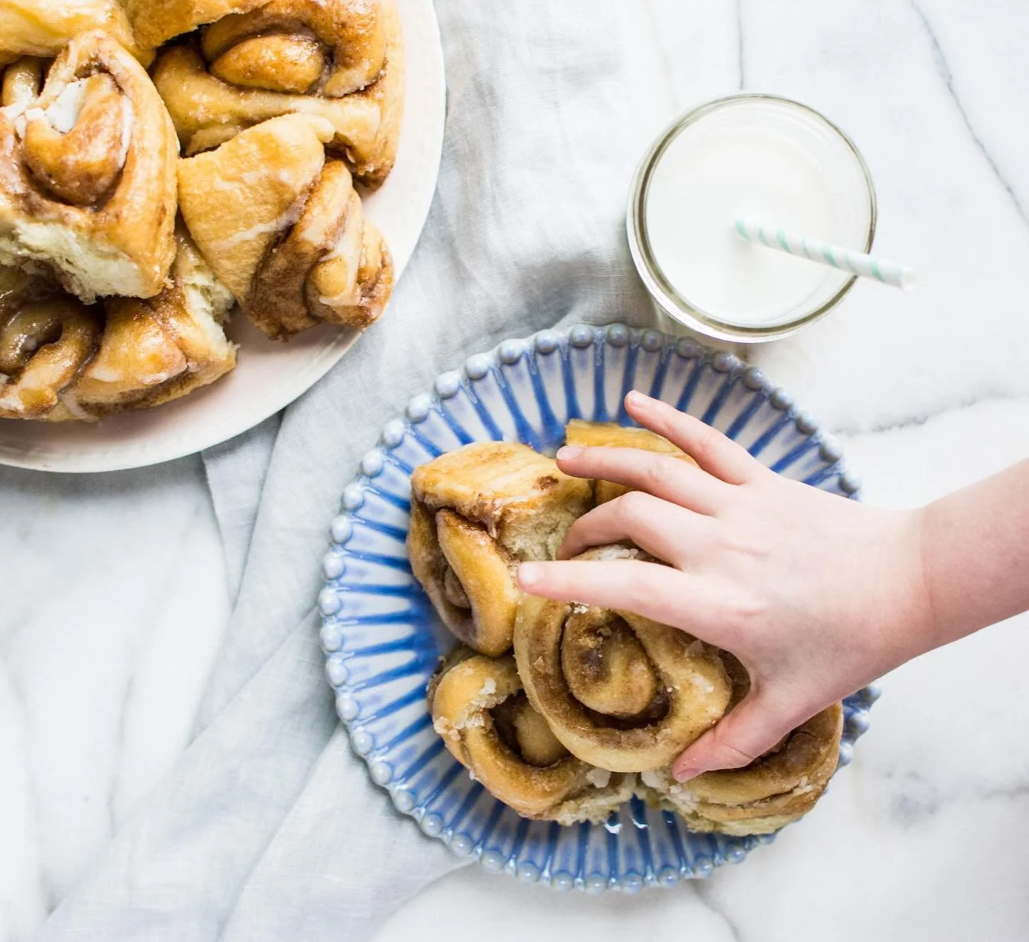 Cinnamon rolls on a blue plate, a cluster of cinnamon rolls on a white plate, a hand reaching for a cinnamon roll, and a glass of milk with a striped straw on a marble surface with a cloth underneath.