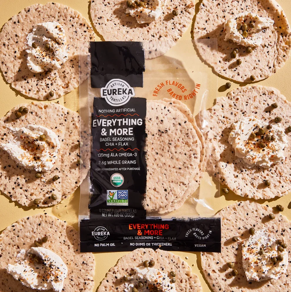 Round, beige, flatbread tortillas garnished with dollops of whipped cream, sprinkled with black and red spices, and topped with small green peppercorns. A package of Eureka artisan flour tortillas labeled "Everything & More" with chia and flax seeds is placed on top of the tortillas. The packaging indicates they are USDA organic, non-GMO, vegan, and contain no palm oil, gums, or thickeners.