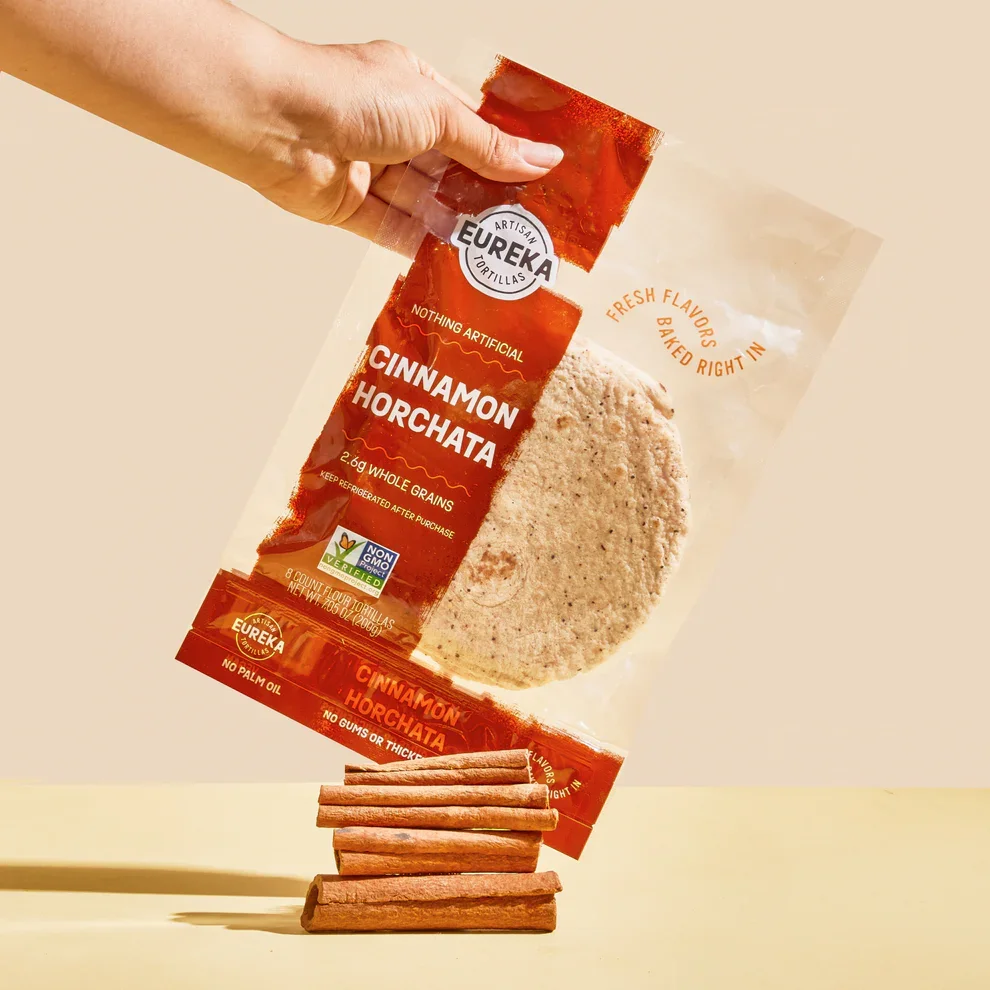 Hand holding a Eureka Cinnamon Horchata tortilla bag with a tortilla partially out of the bag, balanced on a stack of cinnamon sticks.