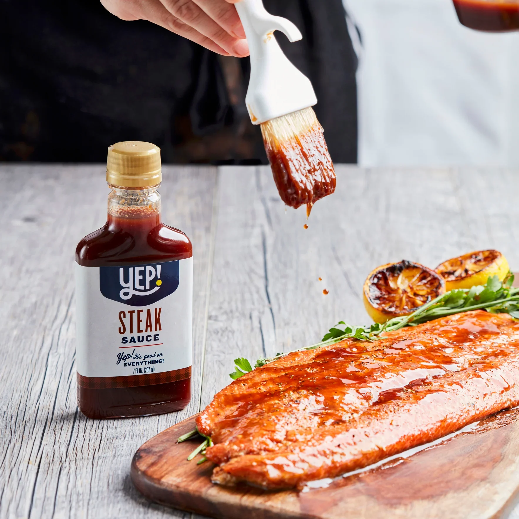A food scene with grilled salmon, grilled vegetables, and a bottle of steak sauce on a white wooden table.