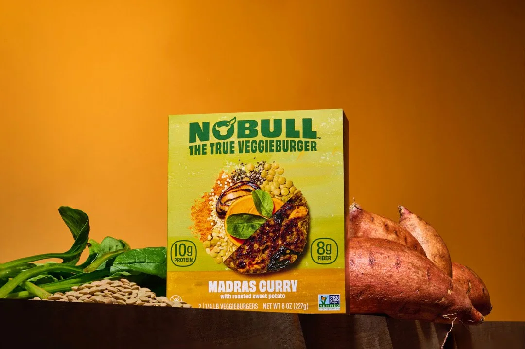 Box of Nobull Madras Curry VeggieBurger with a yellow background, accompanied by sweet potatoes, spinach, sunflower seeds, and lima beans.