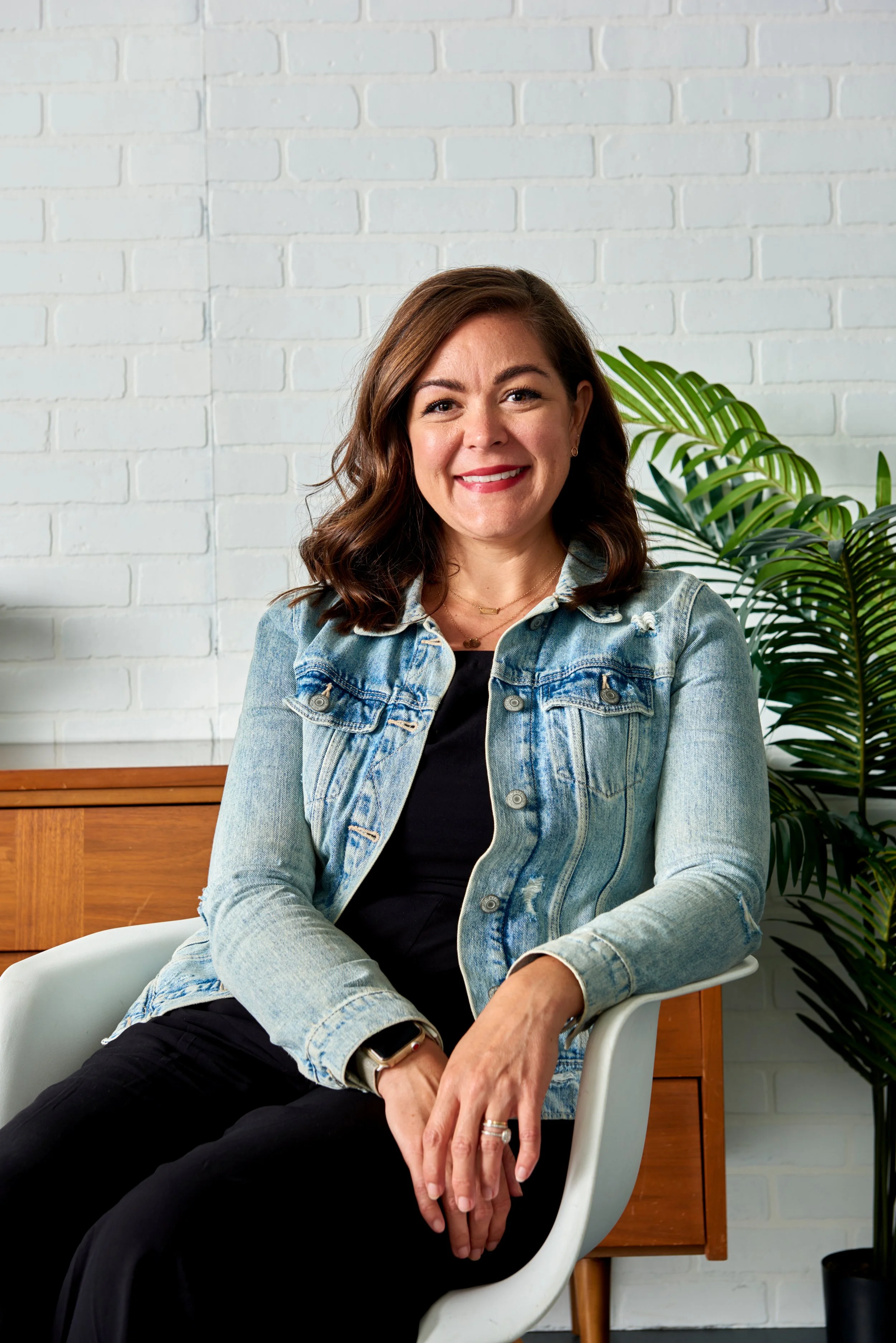 A woman with shoulder-length brown hair, wearing a denim jacket over a black top, sitting on a white chair, smiling, with a large green plant behind her and a white brick wall in the background.