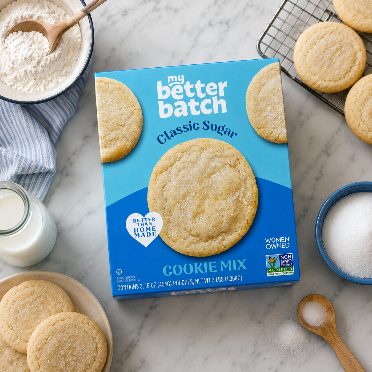 Box of my better batch classic sugar cookie mix on a marble countertop, surrounded by finished cookies, a bowl of sugar, a small jar of milk, and baking utensils.