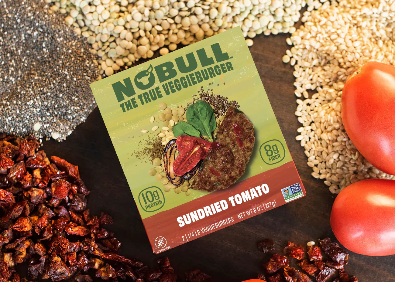 Package of NO BULL veggie burger with sundried tomato flavor, surrounded by sun-dried tomatoes, rice, and seeds on a dark surface.