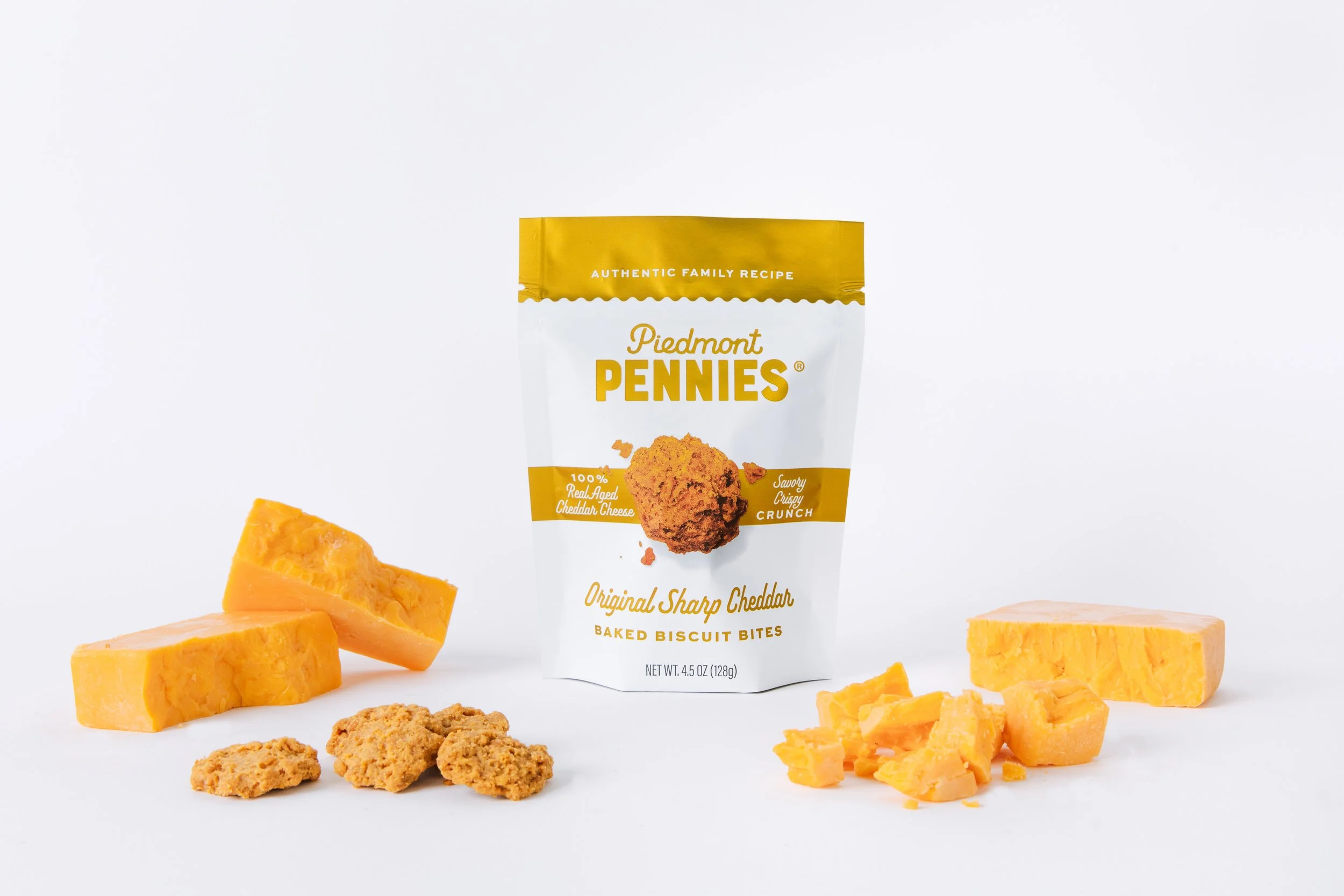 A bag of Piedmont Penny's baked biscuit bites with chunks of cheddar cheese around it on a white background.