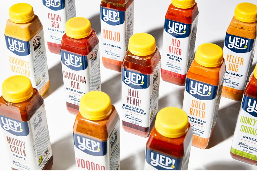 Several bottles of barbecue sauce from the YEP! brand, each with a yellow cap and various labels with different flavors including Cajun Red, Mojo, Carolina Red, Hail Yeah!, Red River, Gold River, Muddy Creek, Voodoo, Honey Sriracha, and Yellow Dog.