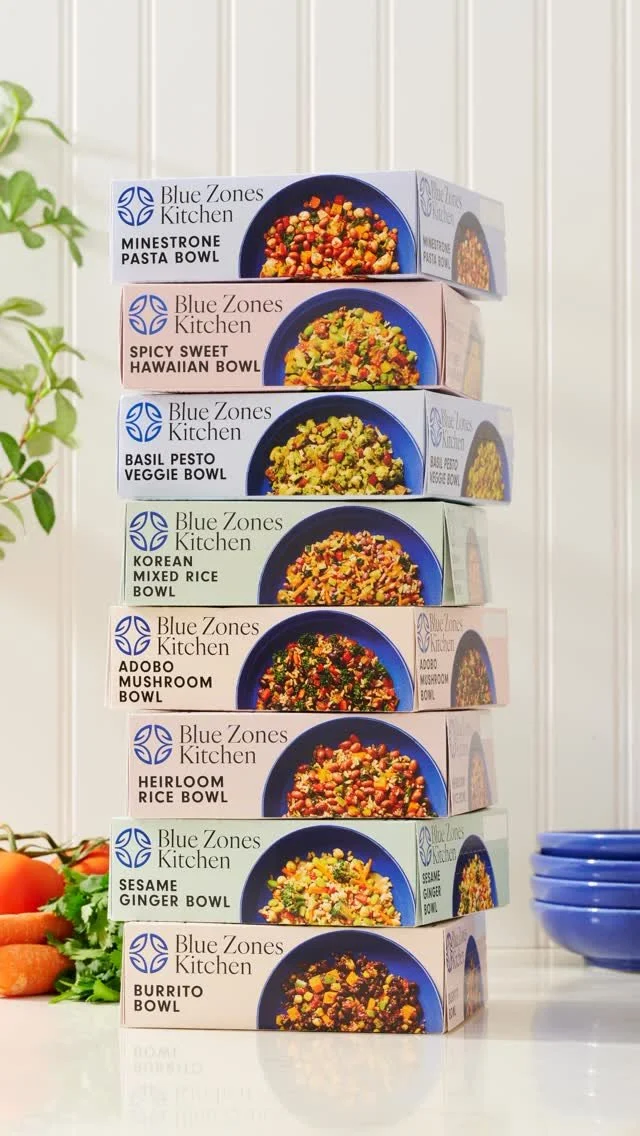 Stacked boxes of Blue Zones Kitchen meal bowls on a white surface with a plant and carrots nearby.