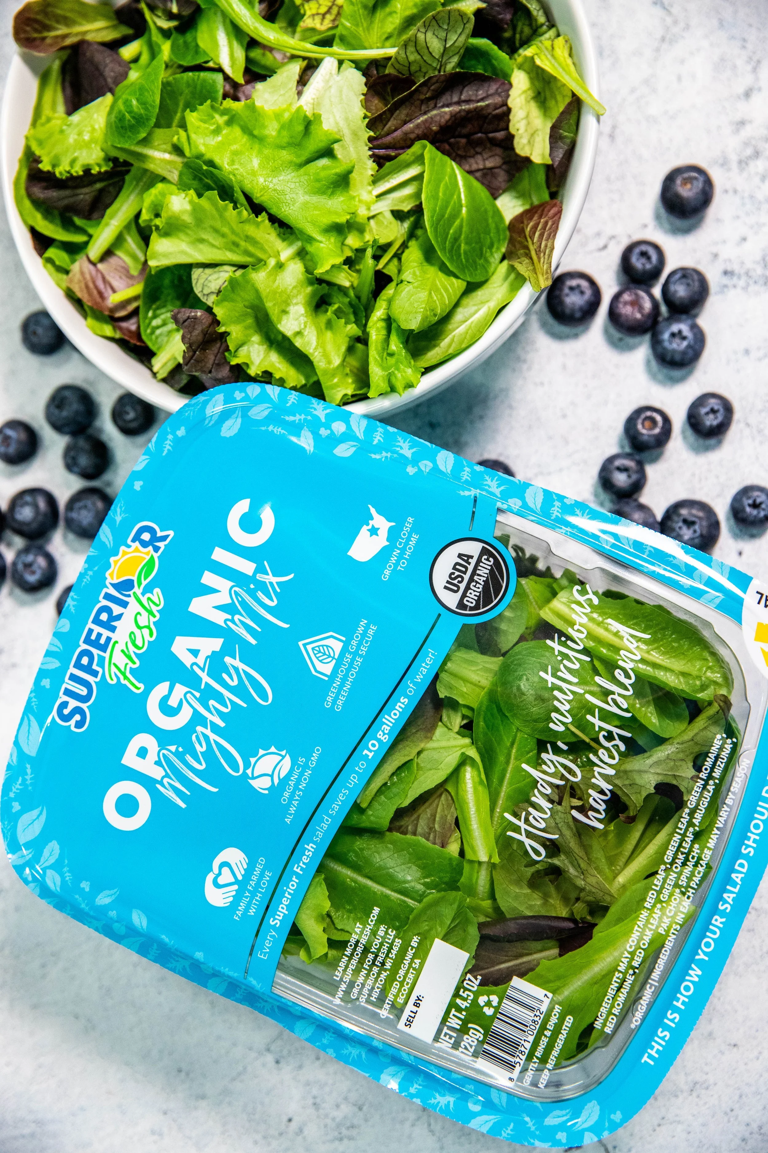 A plastic container of mixed organic salad greens with blueberries scattered around it on a gray surface.