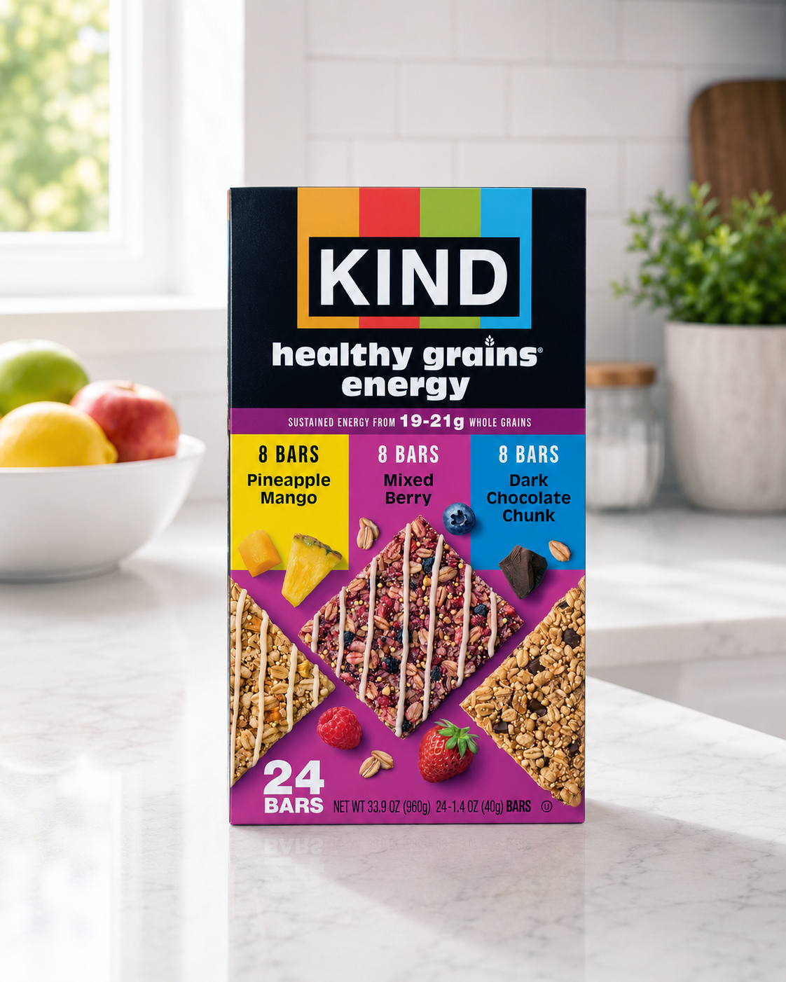 A box of KIND healthy grains energy bars with pineapple mango, mixed berry, and dark chocolate chunk flavors, placed on a kitchen countertop with a bowl of apples and a potted plant in the background.