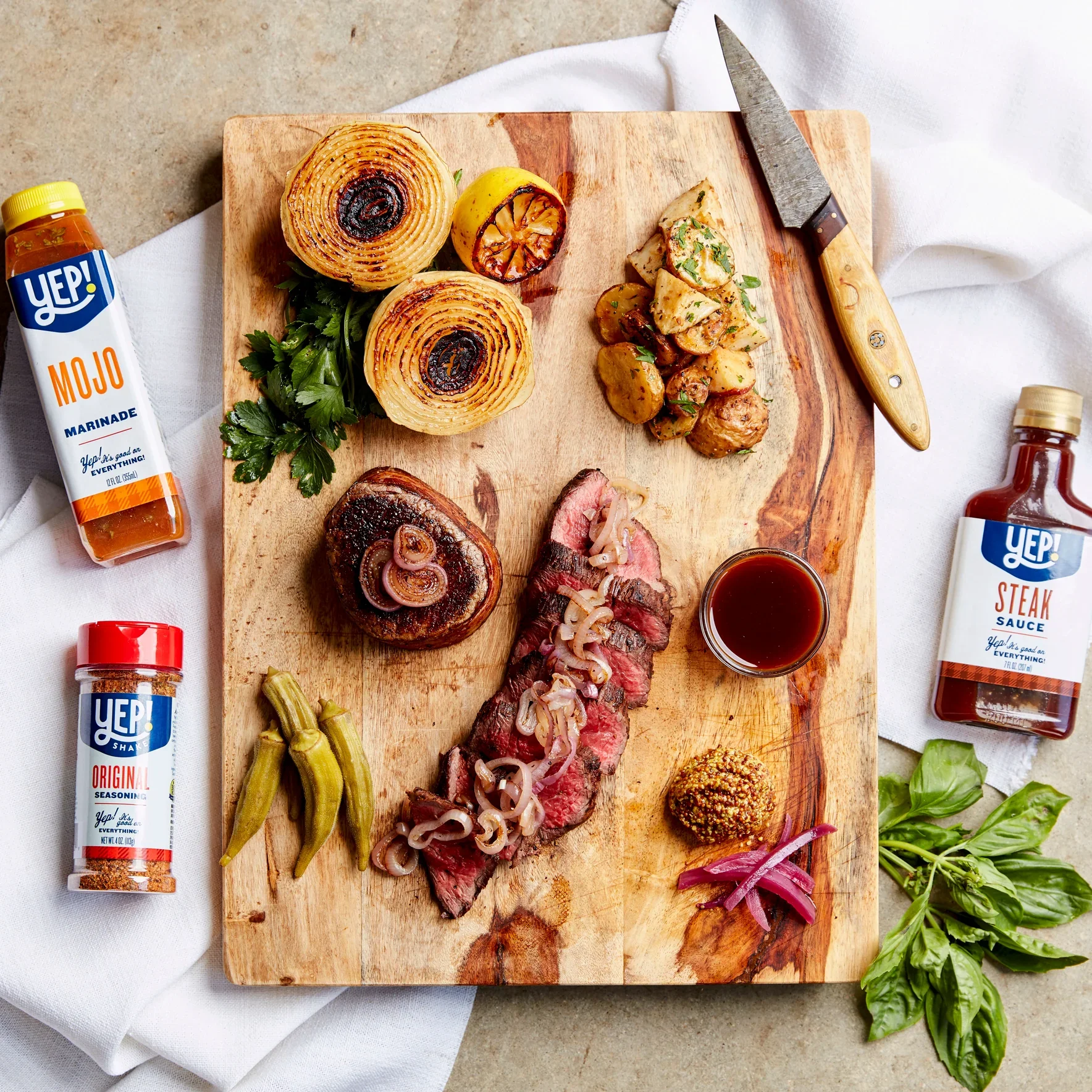 A wooden cutting board on a white cloth with grilled vegetables, sliced steak topped with onions, roasted potatoes, and garnishes. Condiments like marinade, steak sauce, and seasoning bottles are around the board with a knife.