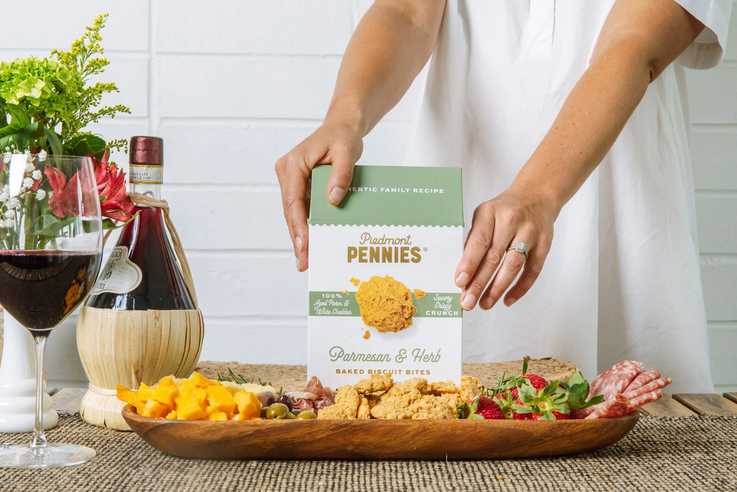 A person holds a box of Piedmont Pennies Parmesan & Herb baked biscuit bites at a table with a food spread, including cheese, grapes, strawberries, charcuterie, and a glass of red wine.