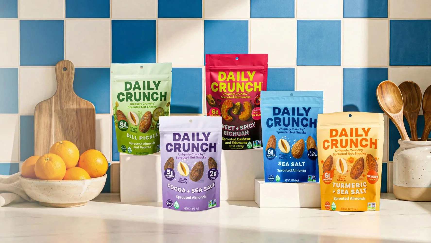 Assorted bags of Daily Crunch sprouted nut snacks on a kitchen countertop with a bowl of lemons and wooden utensils in a white vase. The bags include dill pickle, sea salt, cocoa + sea salt, and turmeric + sea salt flavors, set against a blue and white tiled backsplash.