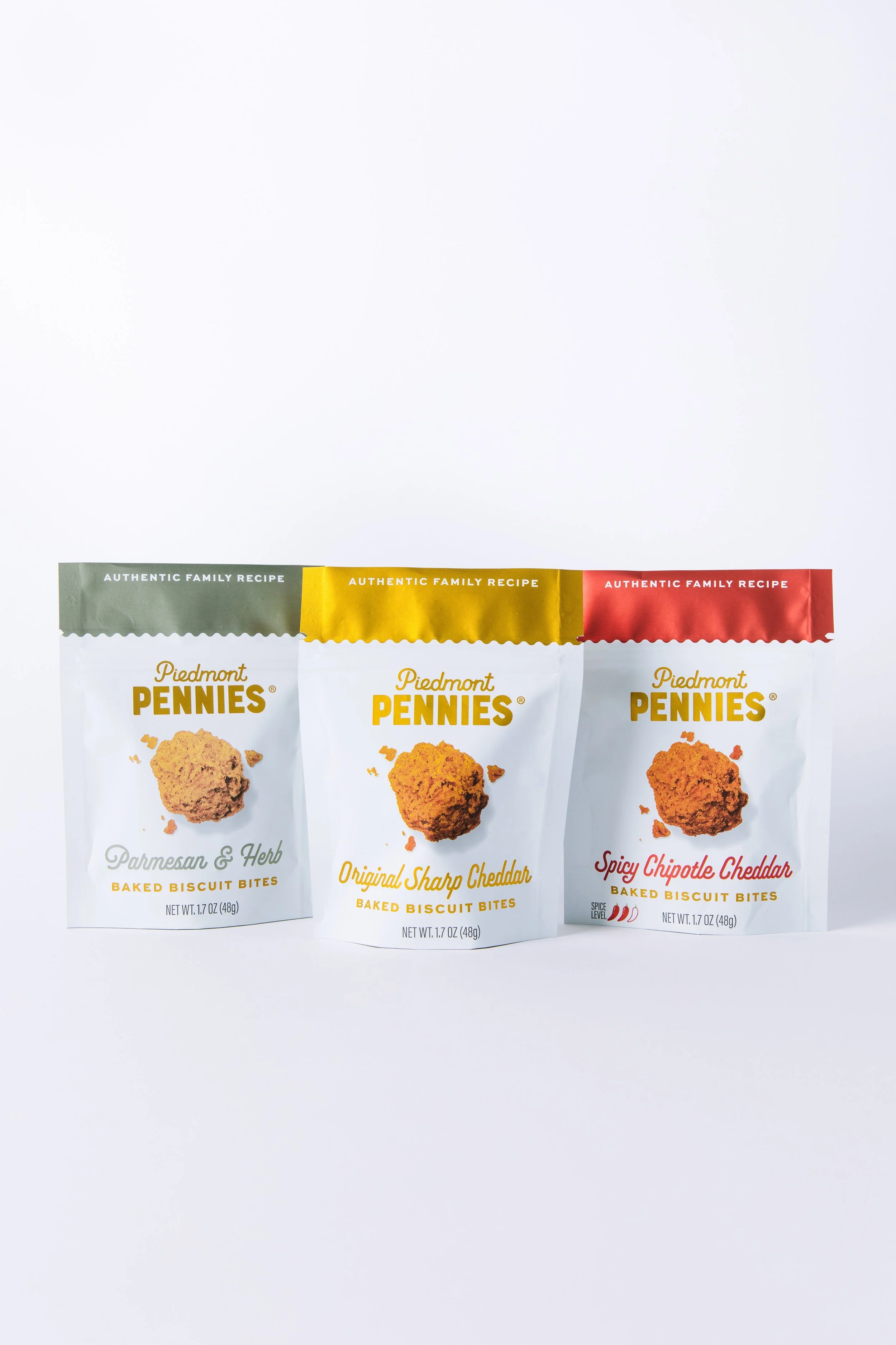 Three bags of Piedmont Pennies baked biscuit bites in flavors Parmesan & Herb, Original Sharp Cheddar, and Spicy Chipotle Cheddar, arranged side by side on a white background.