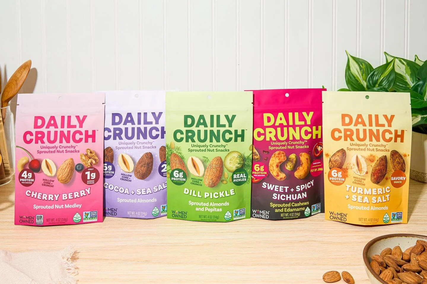 A row of five colorful snack bags on a wooden surface, each featuring different flavors of sprouted nut snacks, with some almonds spilled on the table and a small bowl of almonds nearby.