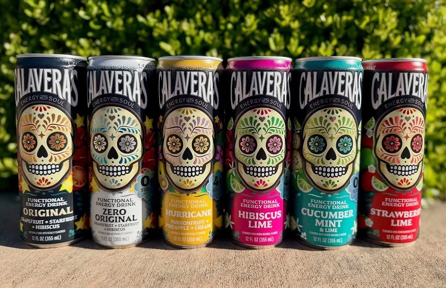 Six cans of CALAVERA'S functional energy drinks in a row, each with a colorful sugar skull design and flavor labels including Original, Zero Original, Hurricane, Hibiscus Lime, Cucumber Mint Lime, and Strawberry Lime, outdoors on a wooden surface with green foliage background.