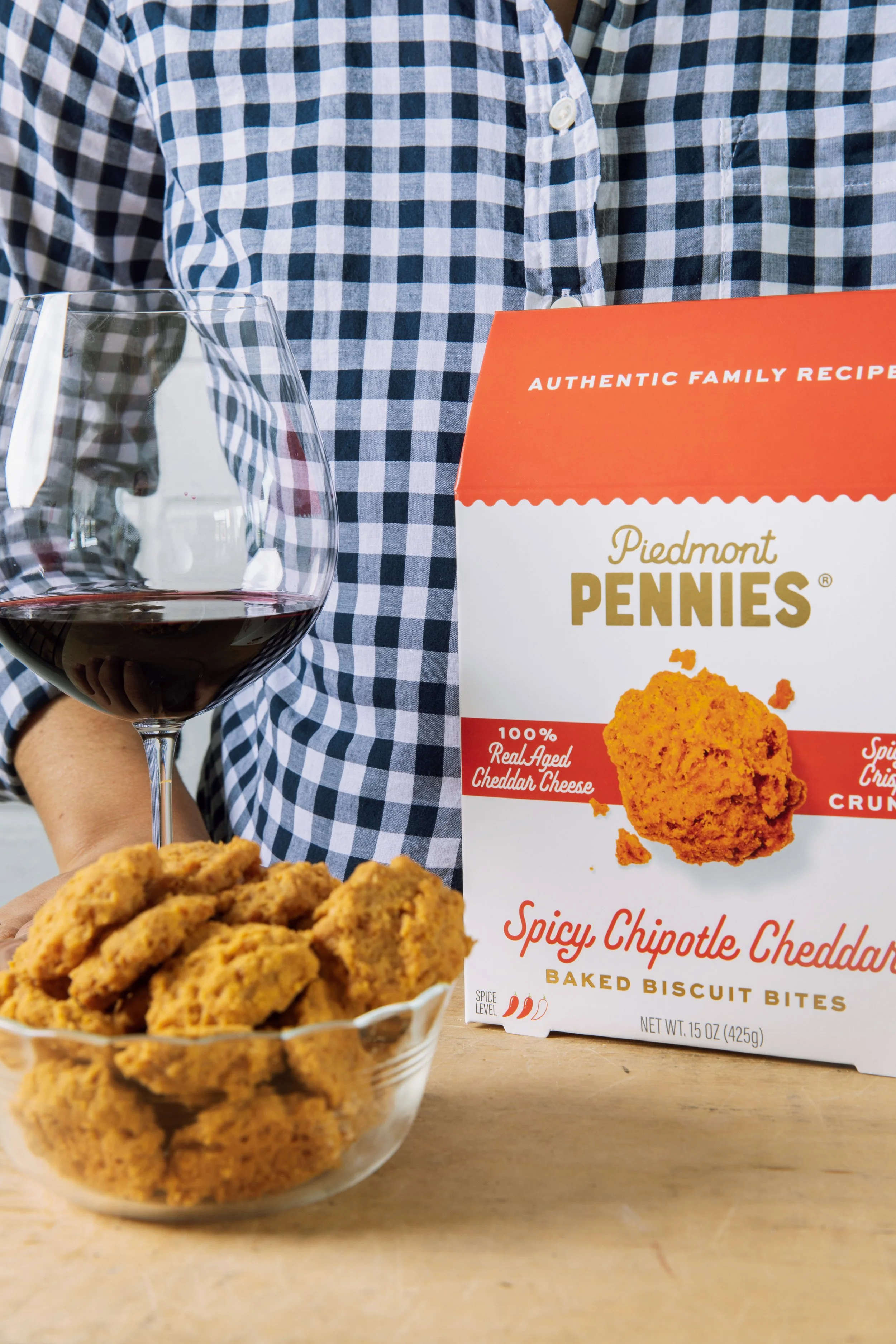 A person wearing a blue and white checkered shirt holding a glass of red wine with a bowl of fried chicken in the foreground and a box of Piedmont Pennies spicy chipotle cheddar baked biscuit bites in the background.