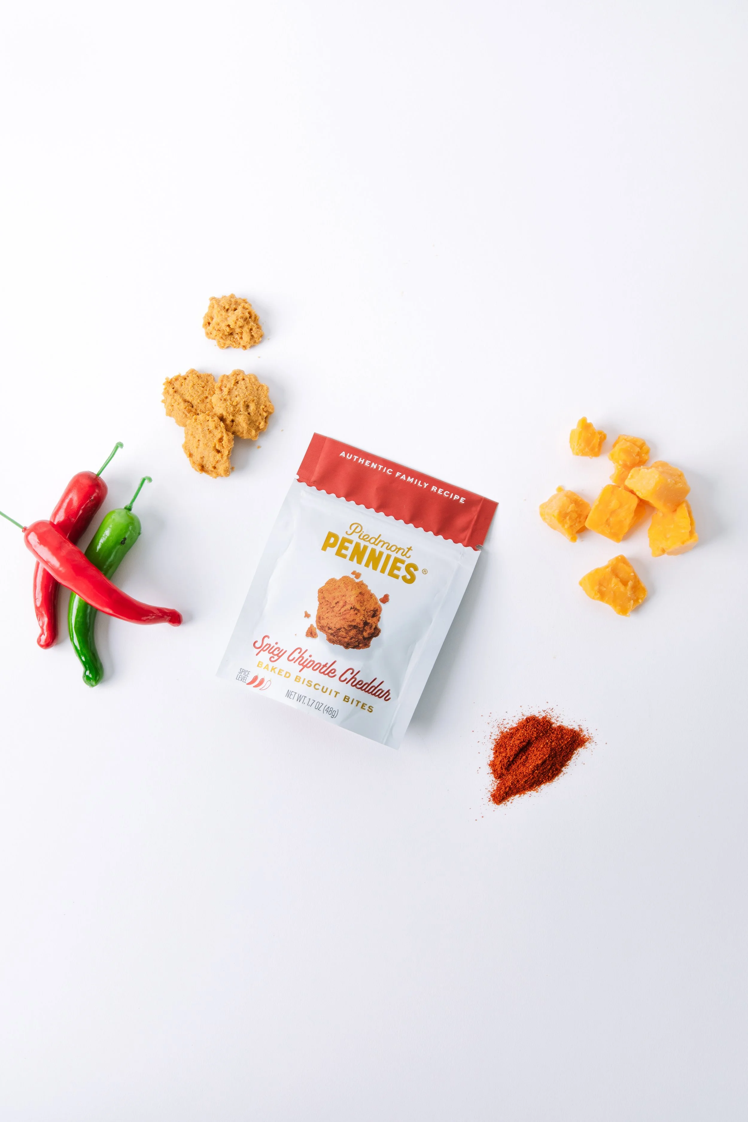 Three red and green chili peppers, a packet of Piedmont Pennies spicy chipotle cheddar baked biscuit bites, two crumbled pieces of cheddar cheese, a pile of red spice, and some broken pieces of baked biscuits on a white background.