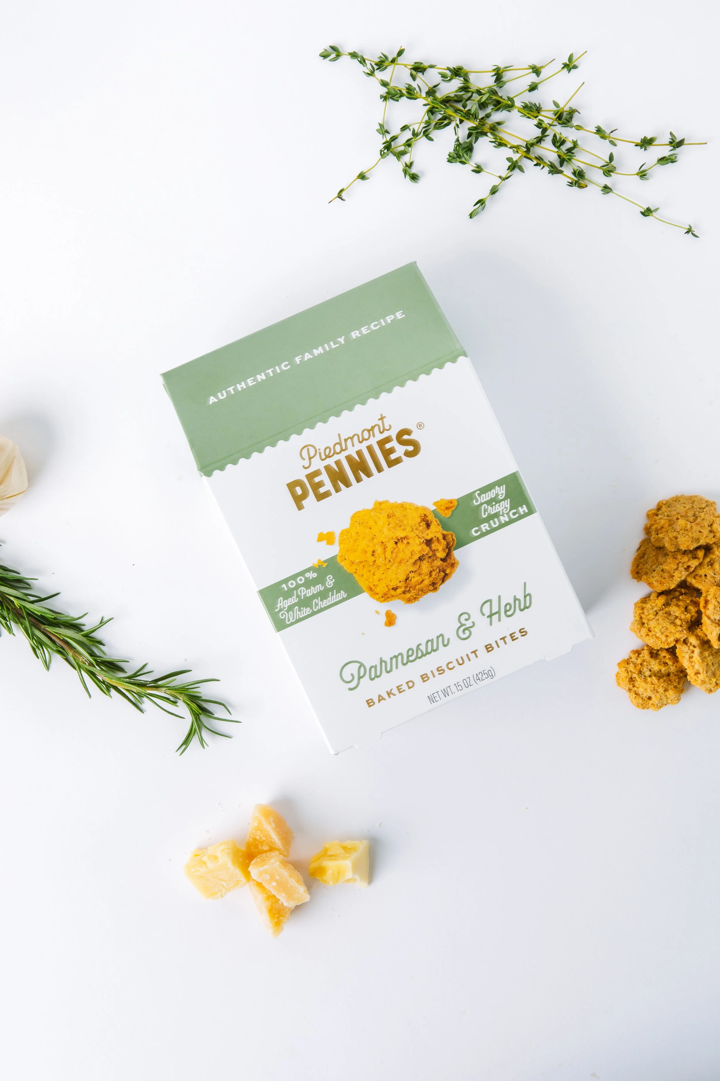 Box of Piedmont Pennies Parmesan and Herb baked biscuit bites on a white background, garnished with sprigs of rosemary and pieces of cheese.
