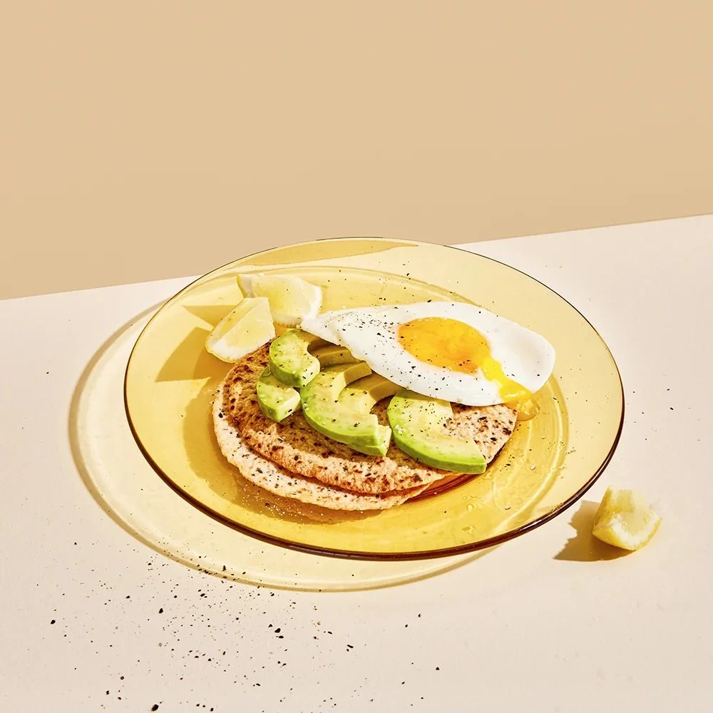 Plate with a fried egg, avocado slices, baked flatbread, and lemon wedges on a yellow plate.