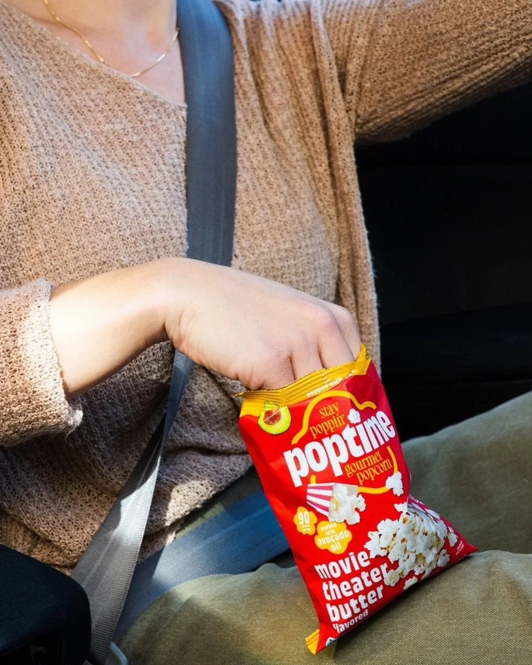 A person reaching into a bag of Popcorn, with the bag labeled 'movie theater butter flavored' and showing an illustration of popcorn and avocado oil, while sitting on a seat with a seatbelt fastened.