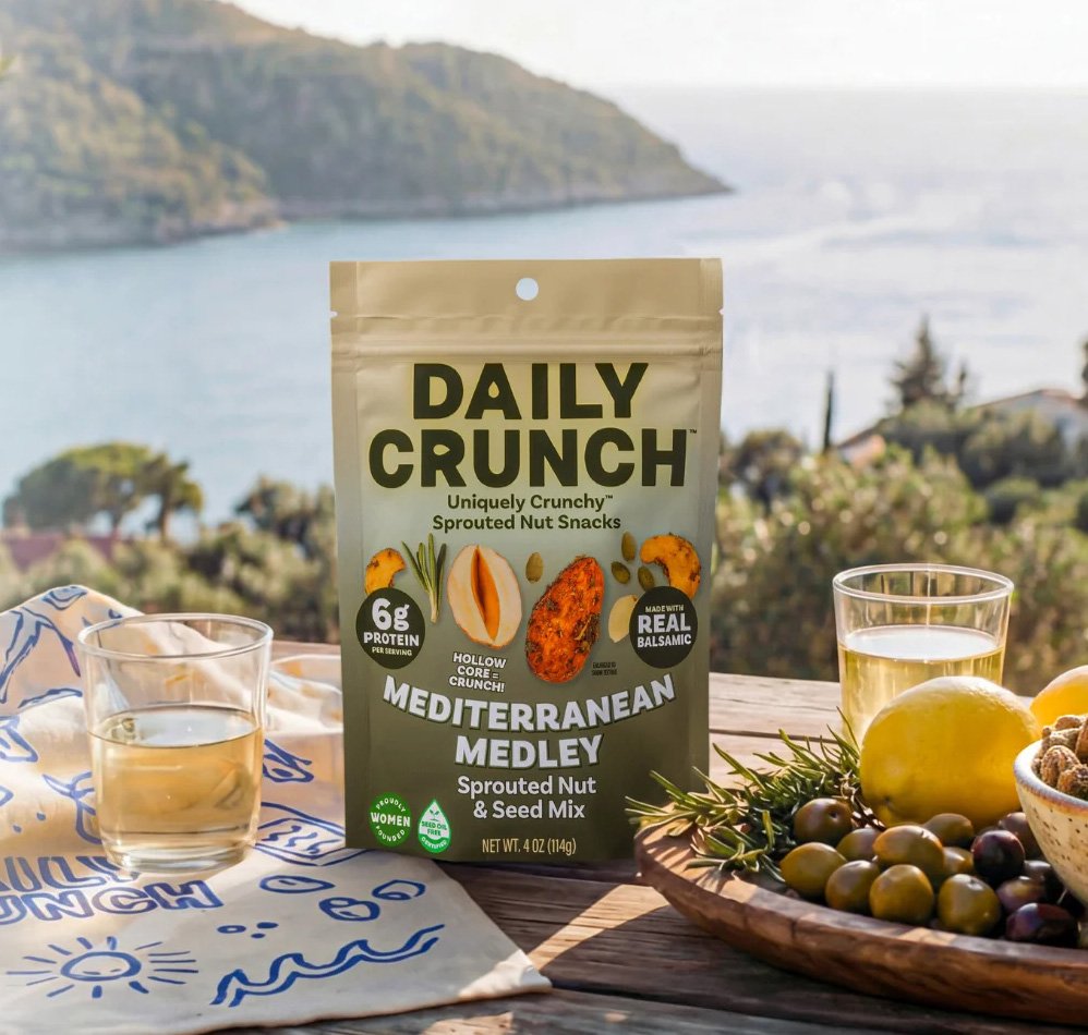 A bag of Daily Crunch Mediterranean Medley sprouted nut and seed mix on an outdoor table, with two glasses of white wine and a bowl of assorted fruit and nuts, overlooking a scenic body of water and hills in the background.
