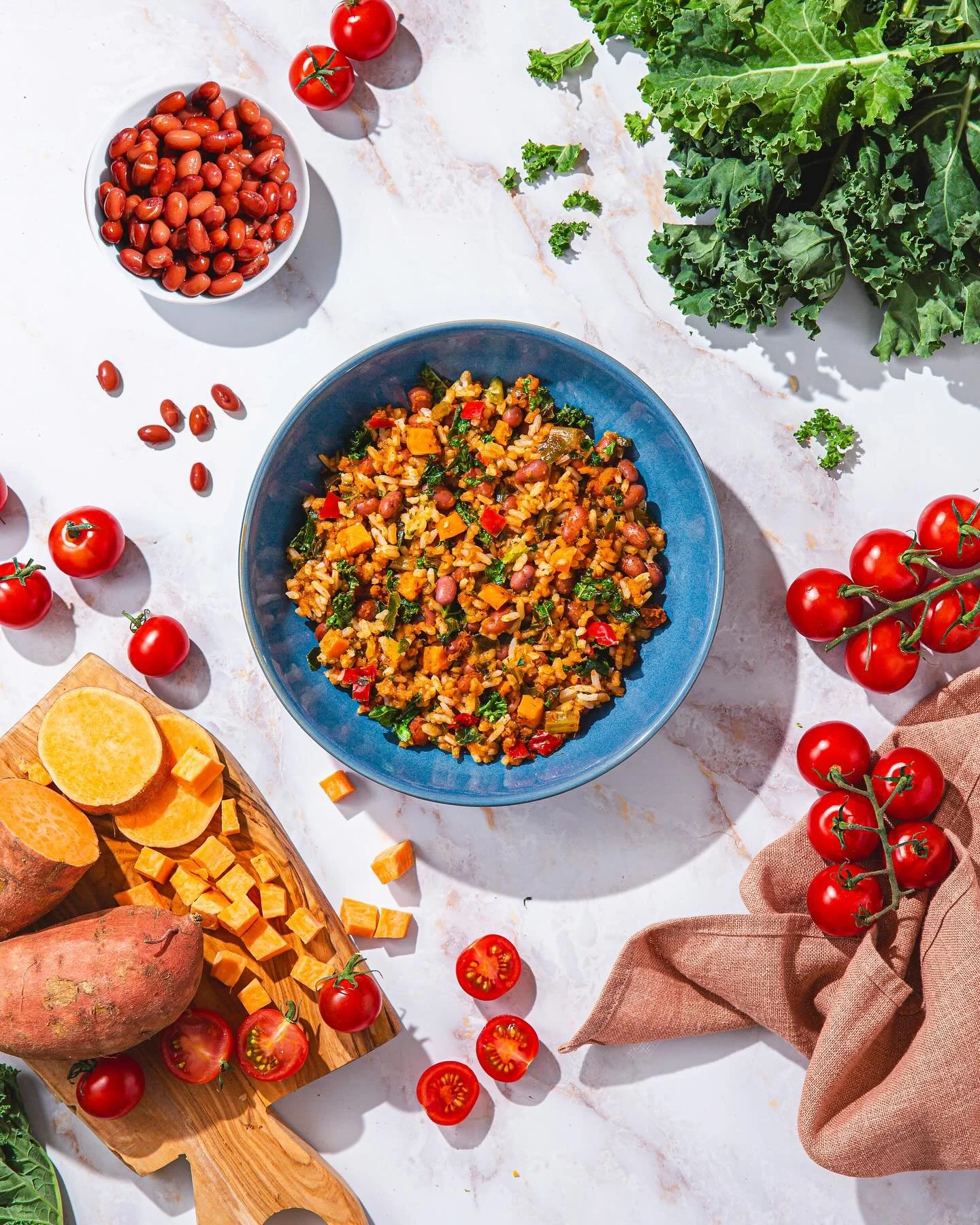 A top-down photo of a bowl of cooked rice and beans with vegetables, surrounded by fresh ingredients including cherry tomatoes, sweet potatoes, and kale on a light-colored surface.