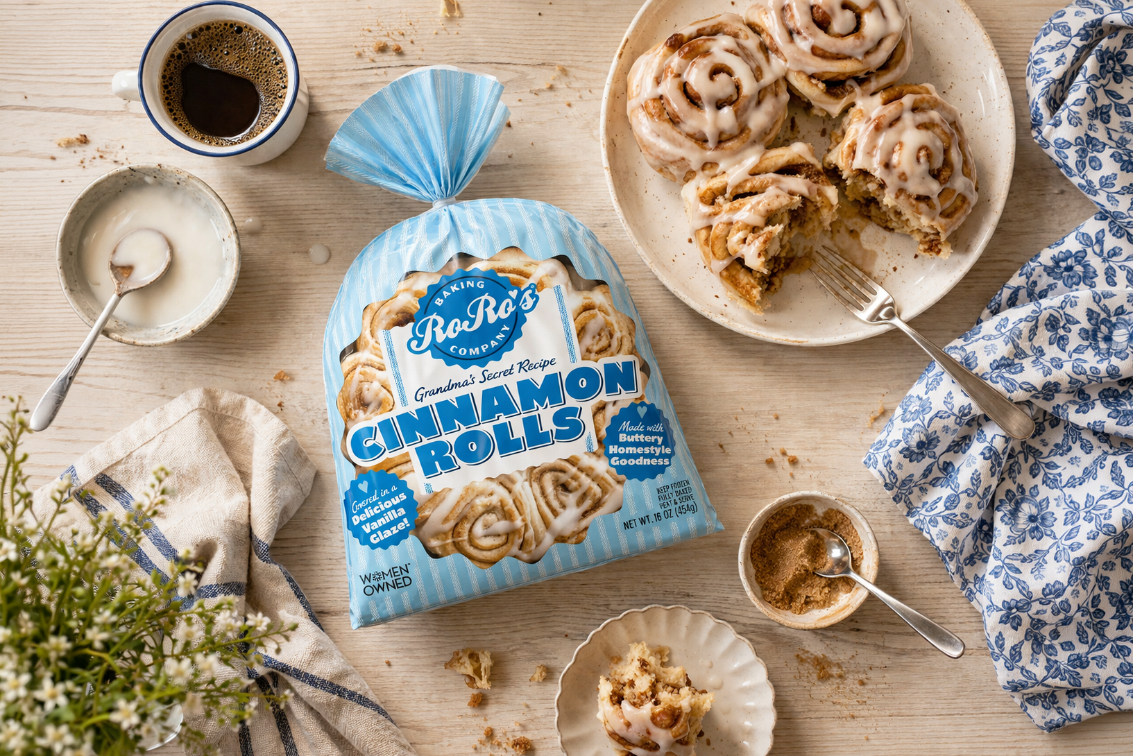 A package of Ra-Ra's Cinnamon Rolls with a plate of cinnamon rolls topped with icing, a cup of coffee, and small bowls of icing and cinnamon sugar on a wooden table with a blue patterned cloth and a small plant.
